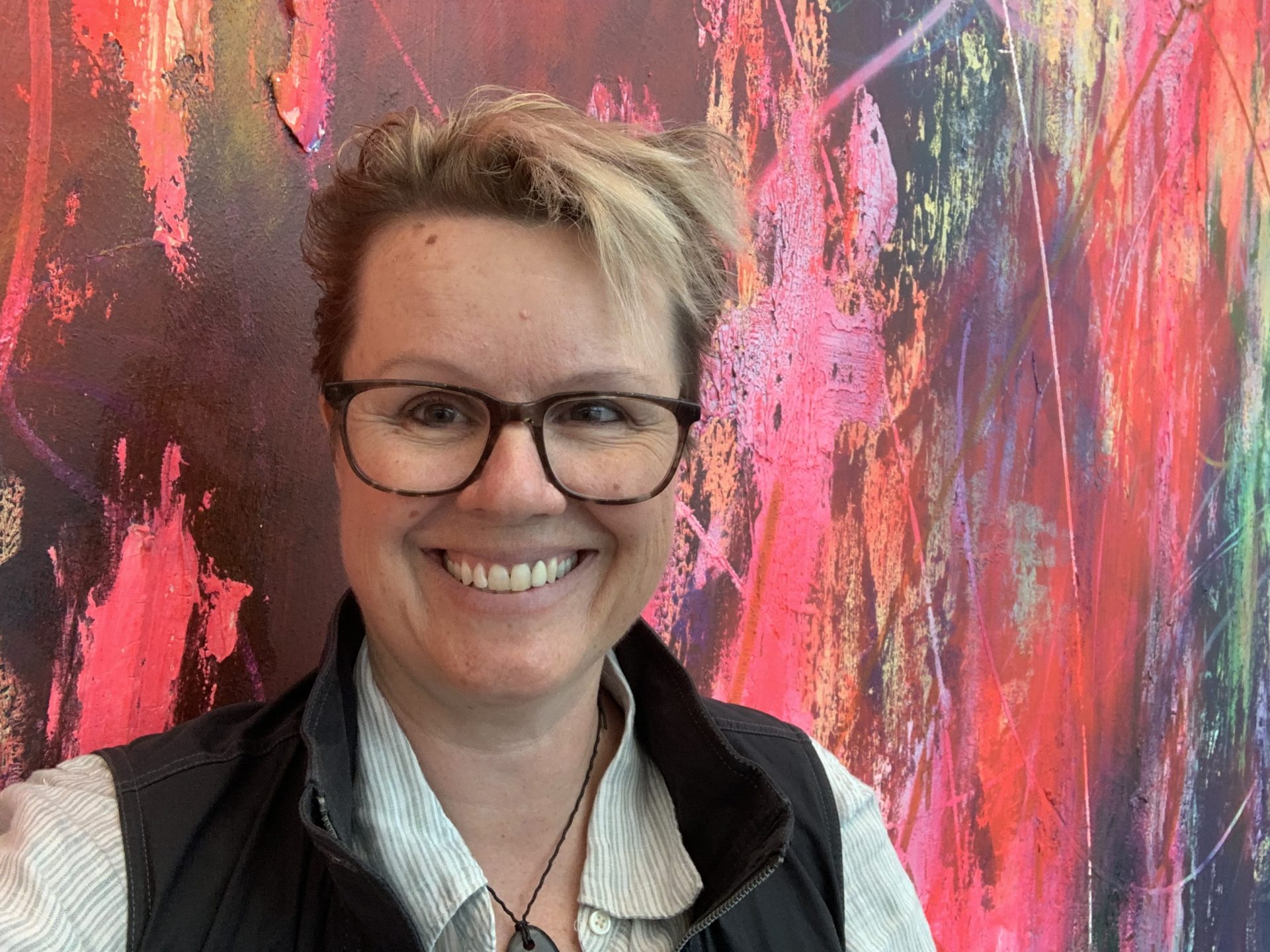 Smiling woman with short blond hair and glasses against a colorful backdrop
