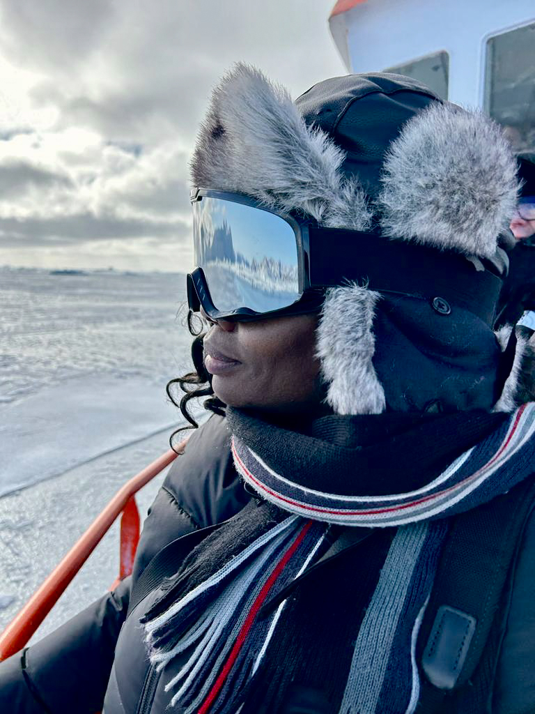 A woman in a fur-lined hat and snow goggles stands on the deck of a shop and looks out of frame as she sails through icy waters.