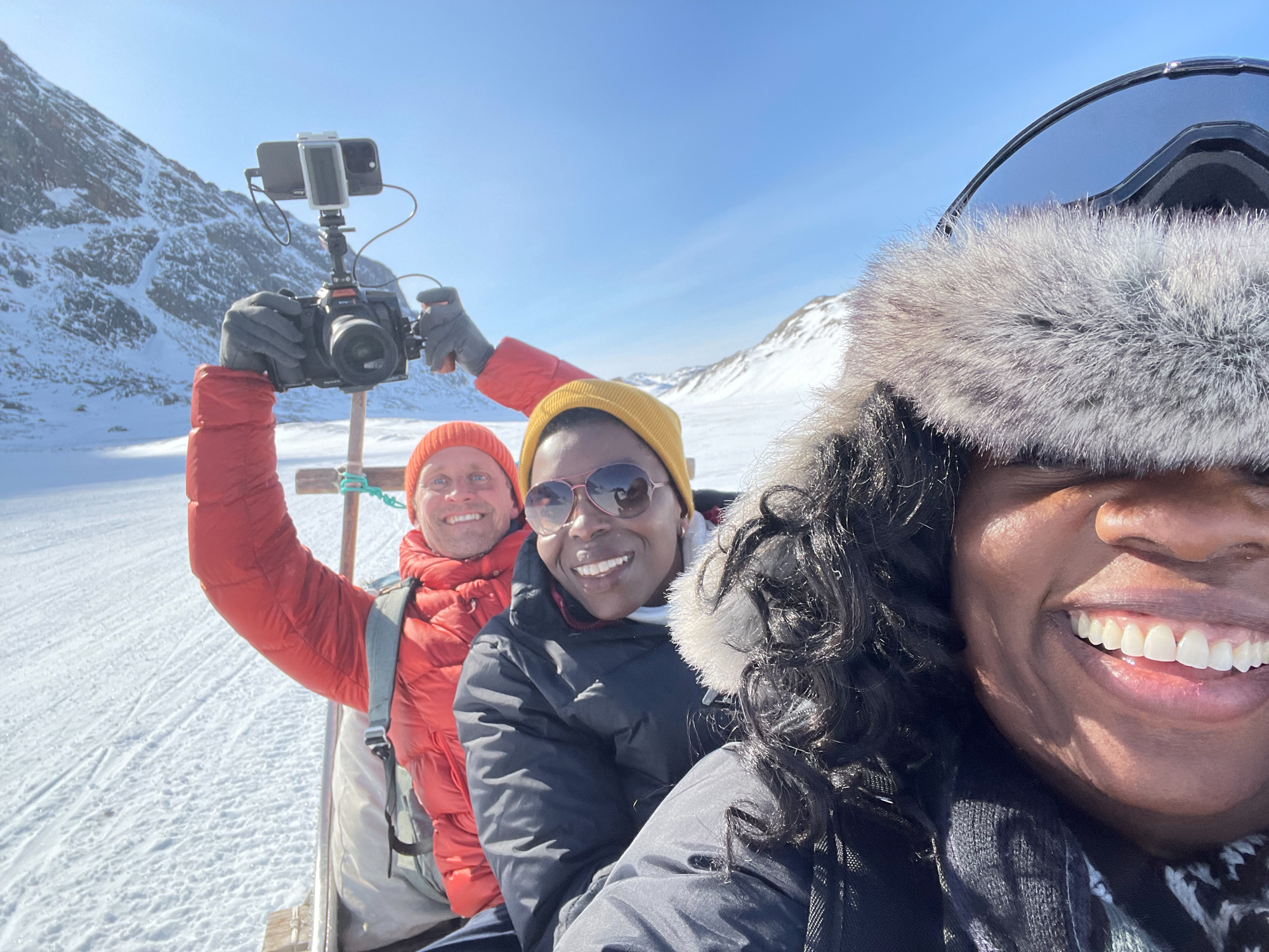 Three smiling people — two women and one man, on the back of a sled in a snowy landscape.