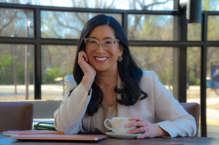 Portrait of a seated woman with black hair and glasses in a suit, holding a ceramic coffee cup.