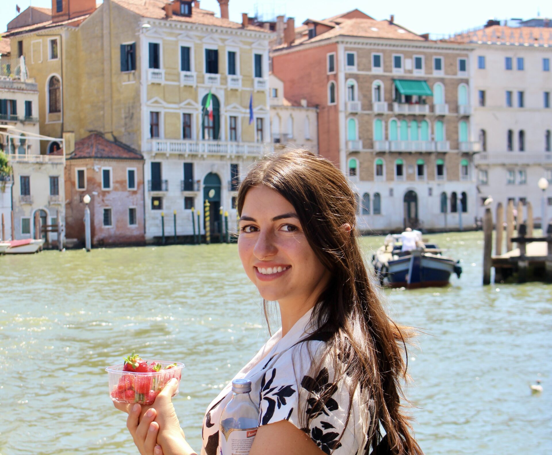 Woman with long hair holding a container of strawberries in front of a canal with a rowboat and old buildings in the background.