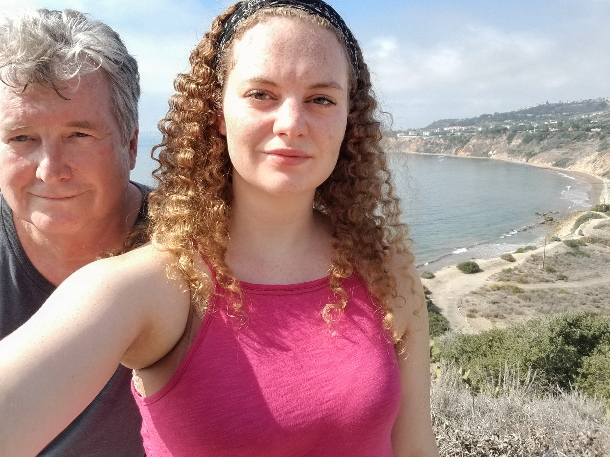 Selfie of a young woman with curly hair and her father with a sandy cove and ocean in the background.