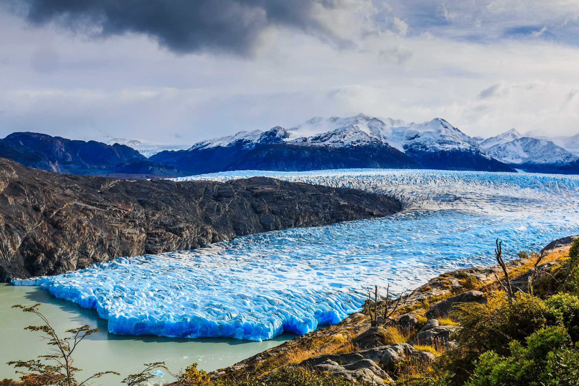 Large glacier in dappled sunshine and clouds with snow-capped mountains in the background.
