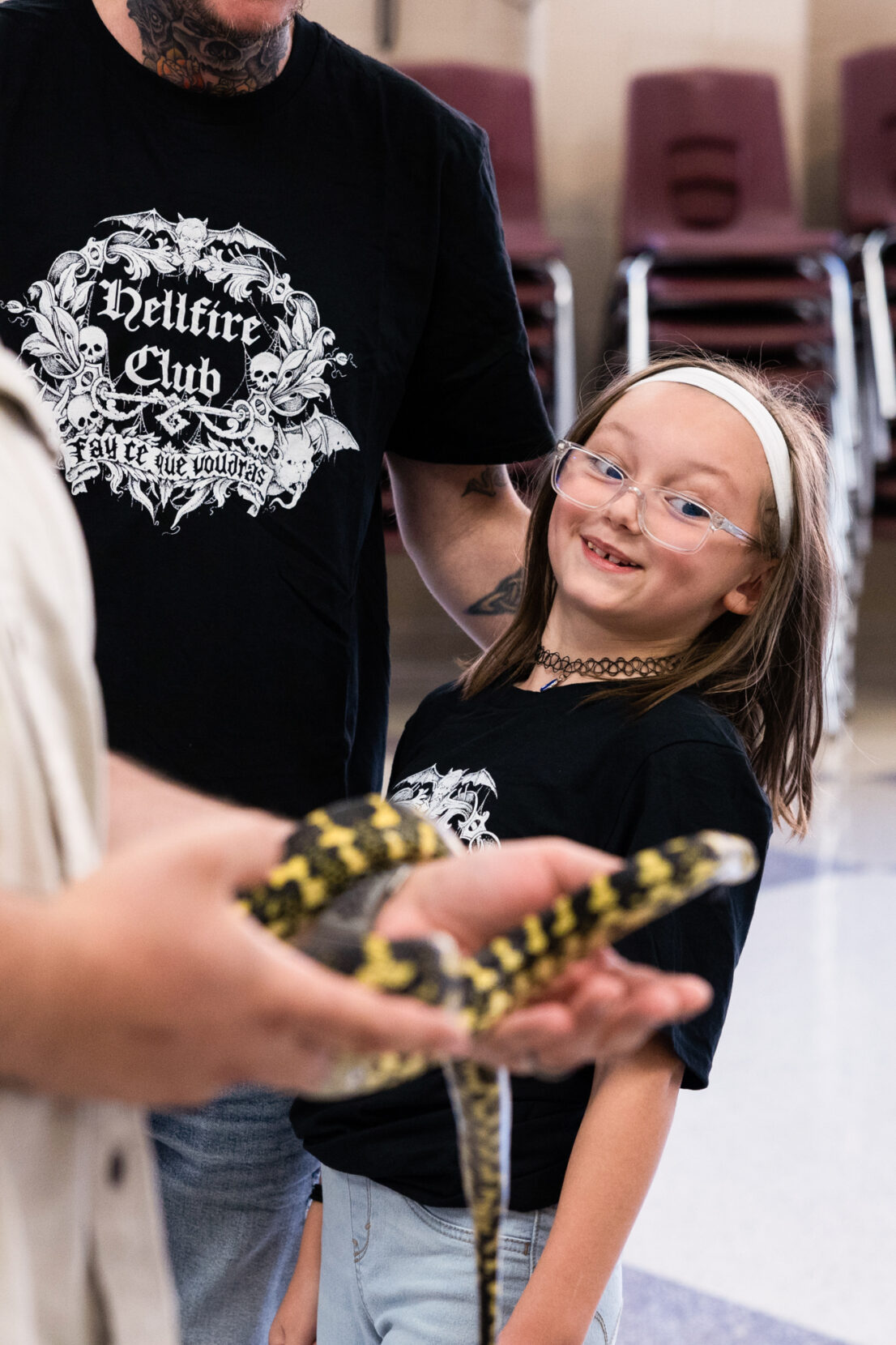 A young girl in a white headband and glasses smiles at a snake being held in the foreground.