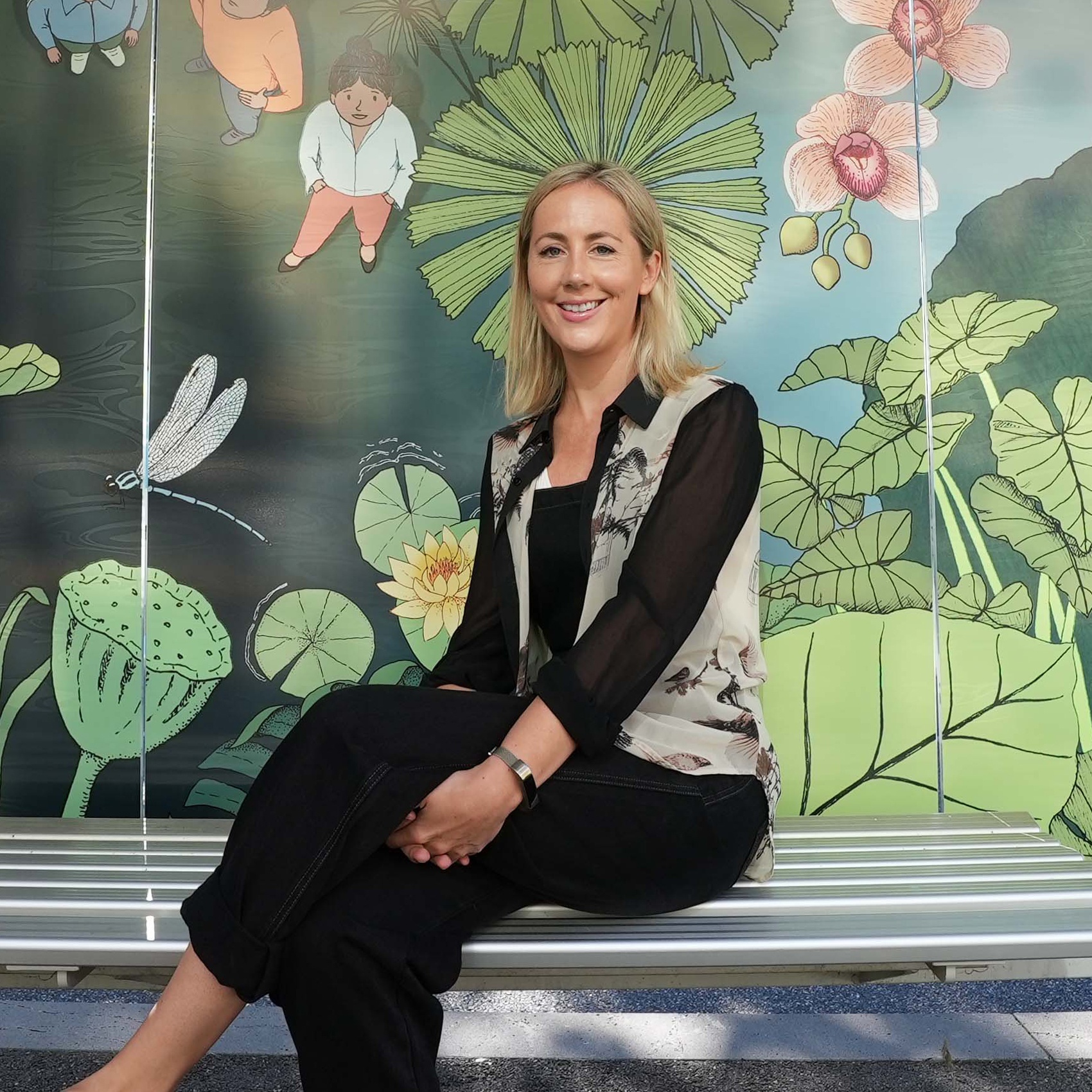 Portrait of a woman sitting on a bench in front of a mural depicting plants and flowers.