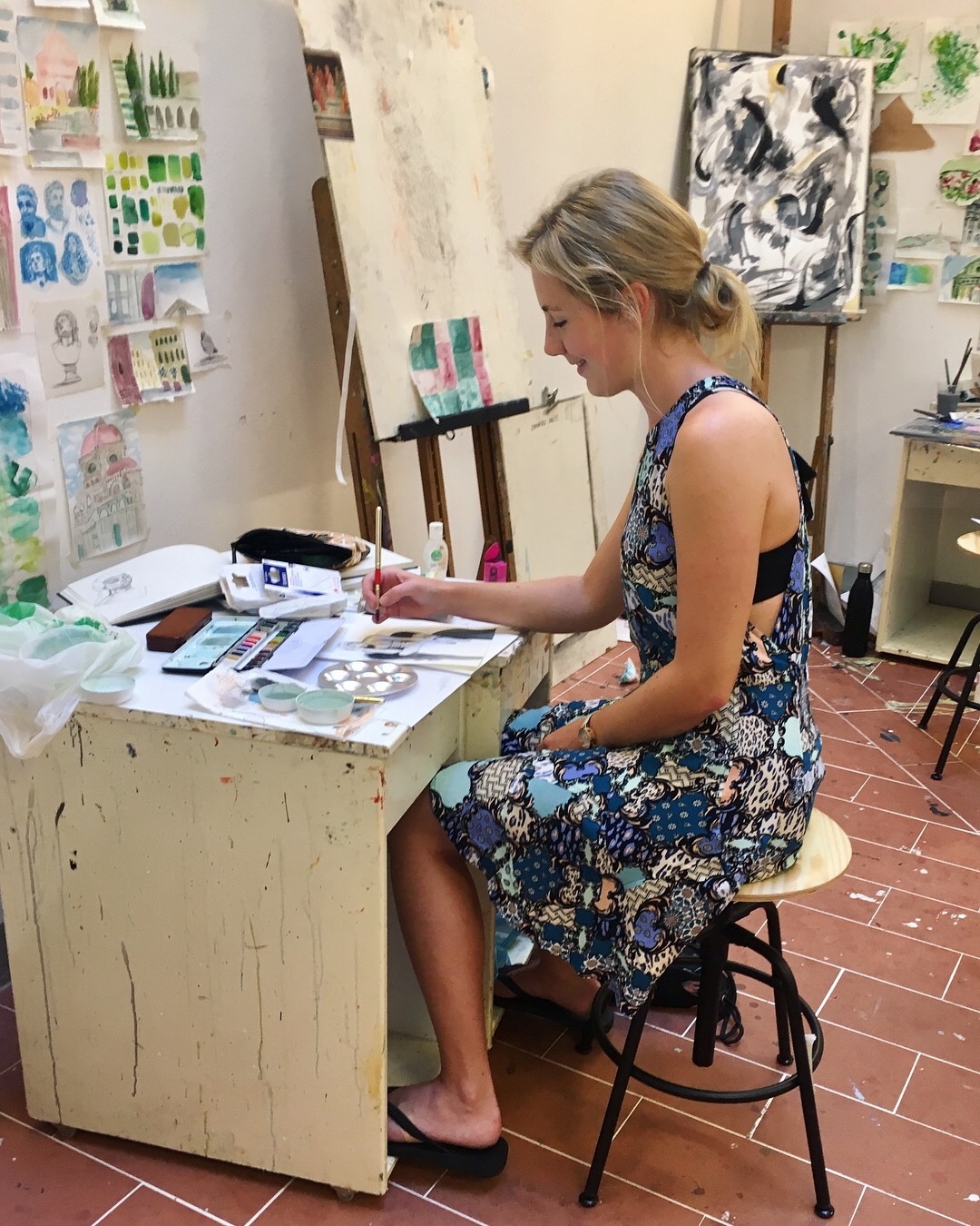 Blonde woman in a dress sitting at a desk, painting.