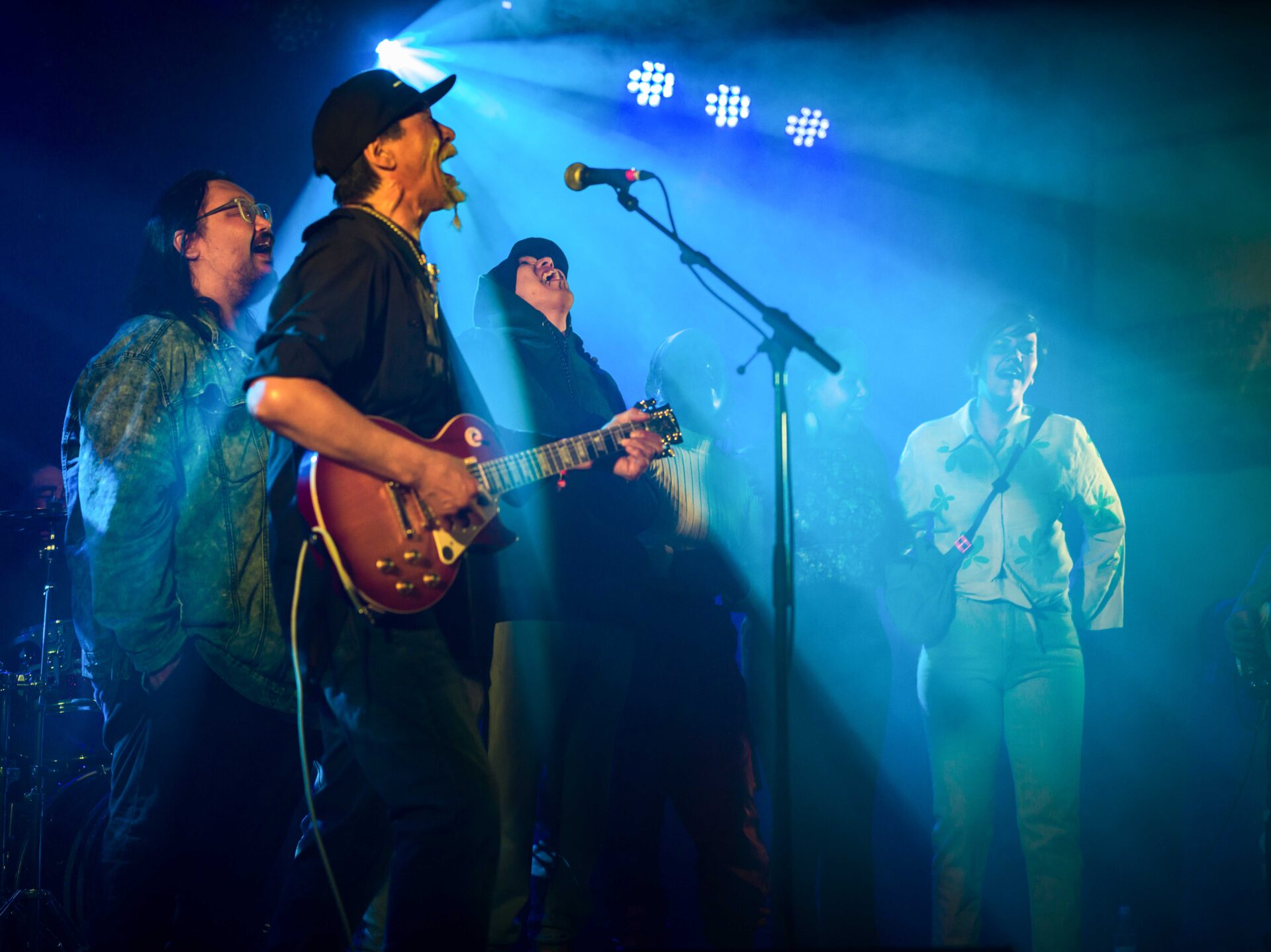 Four singing musicians in hoodies and denim jackets stand backlit with blue and white stage lights before a microphone, mouths open in song.