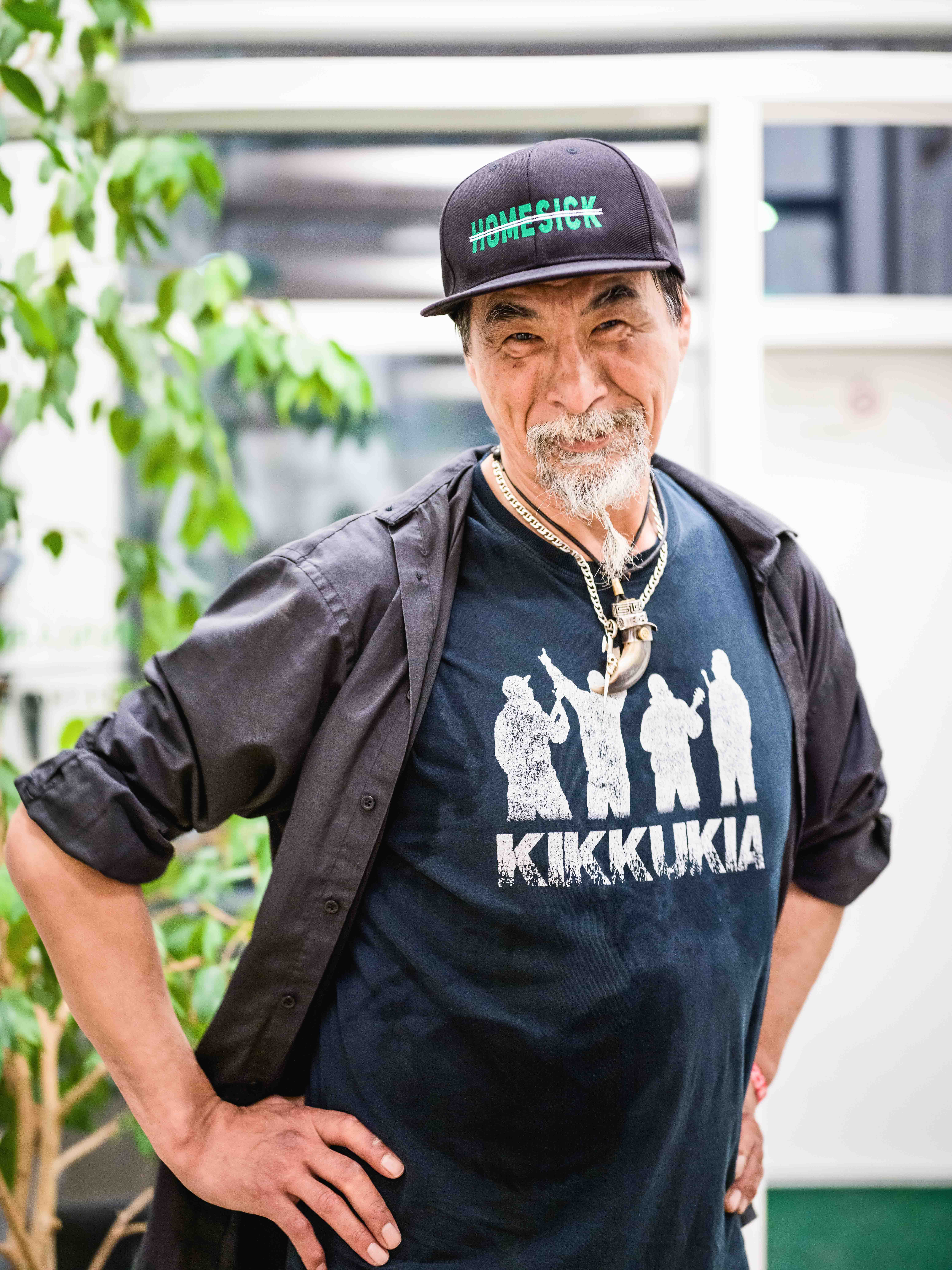An older man man with a white goatee and dark eyebrows smiles at the camera while wearing a baseball cap with the word Homesick on it, and a dark blue-green t-shirt that reads Kikkukia. His hands are on his hips.