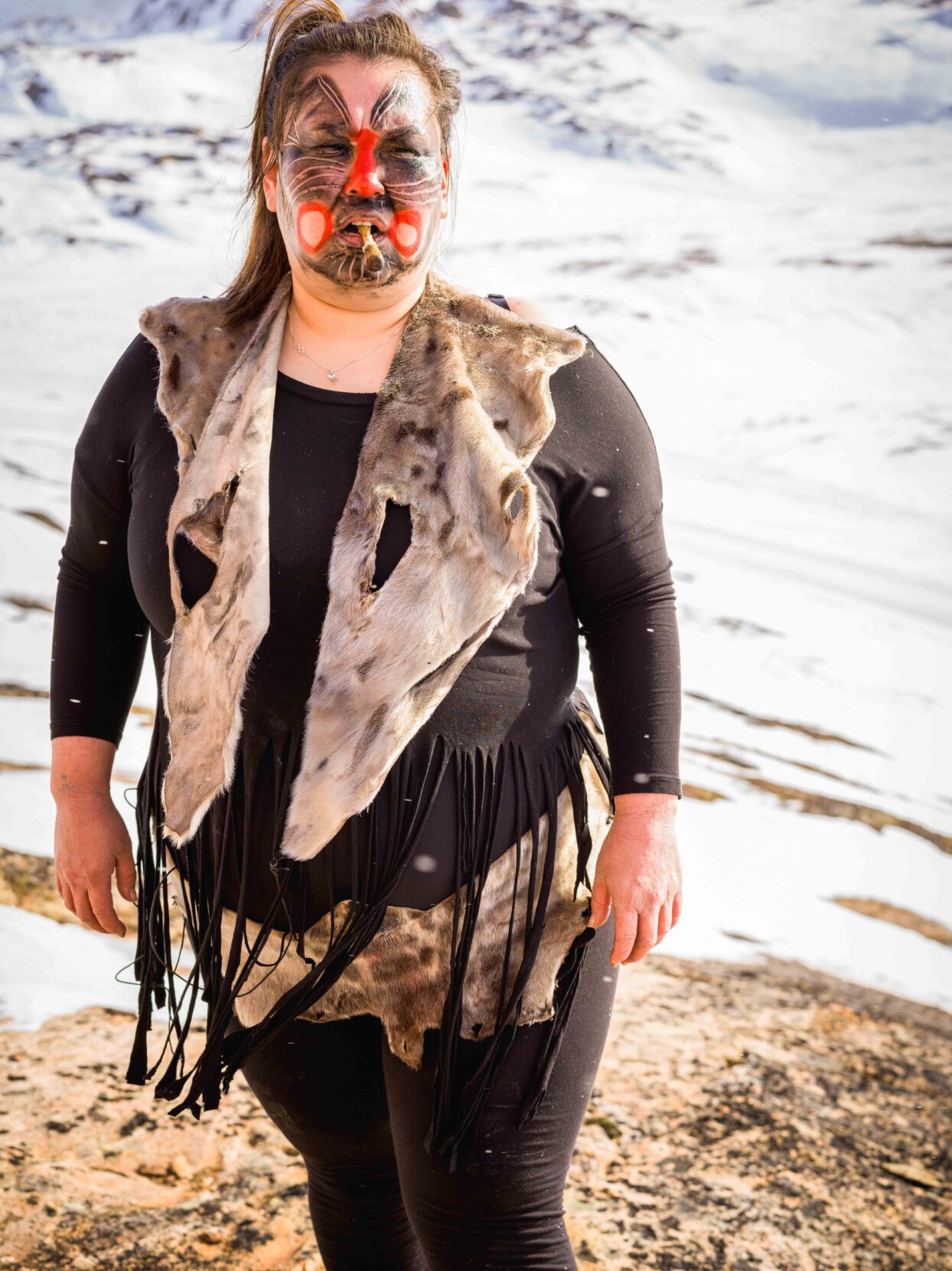 A woman wearing black clothing and animal skins with red and black face paint stands before a snowy landscape.