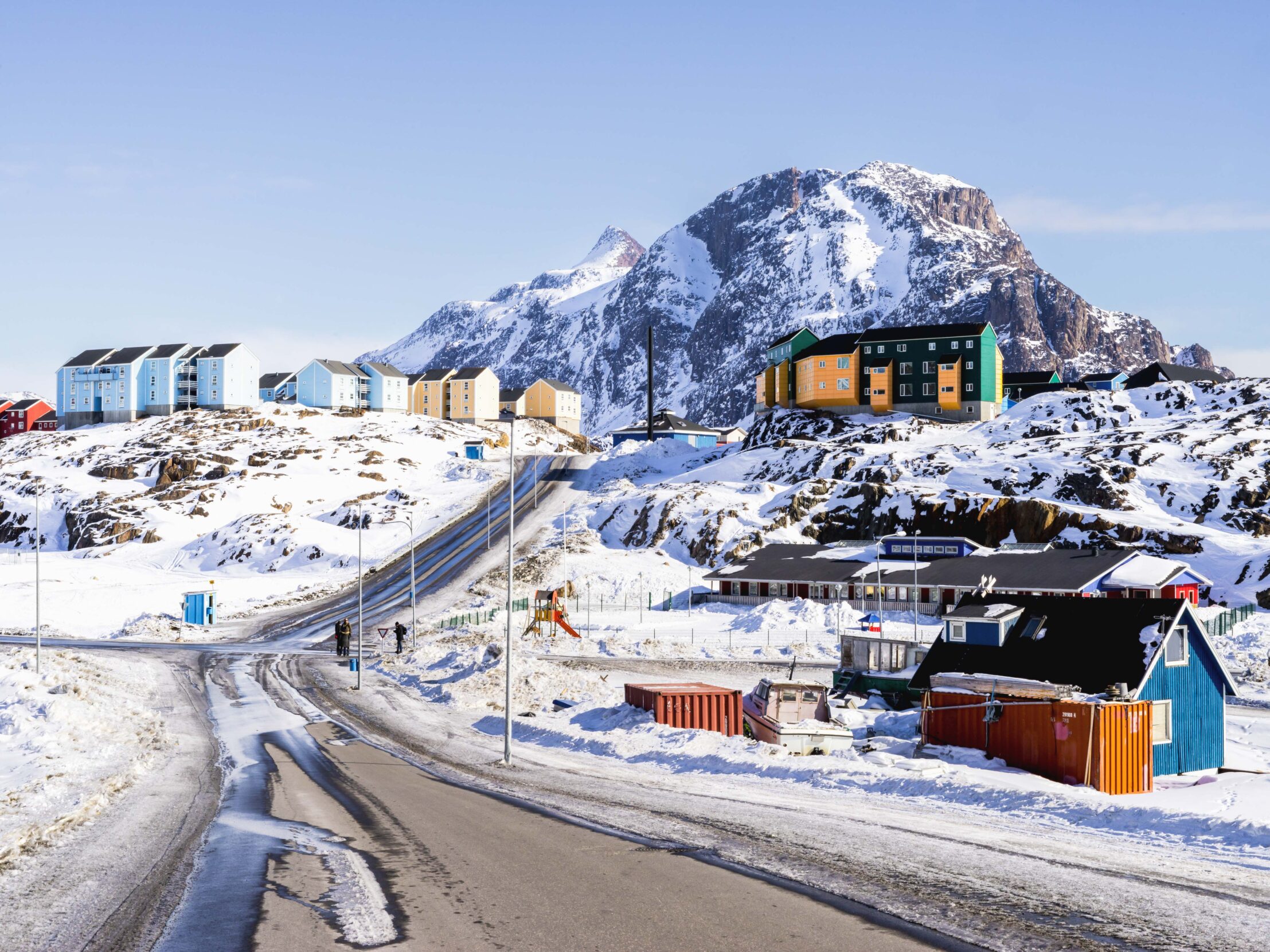 A paved road in a sunny, snowy town of brightly colored buildings leads to a rocky mountain dusted with snow.