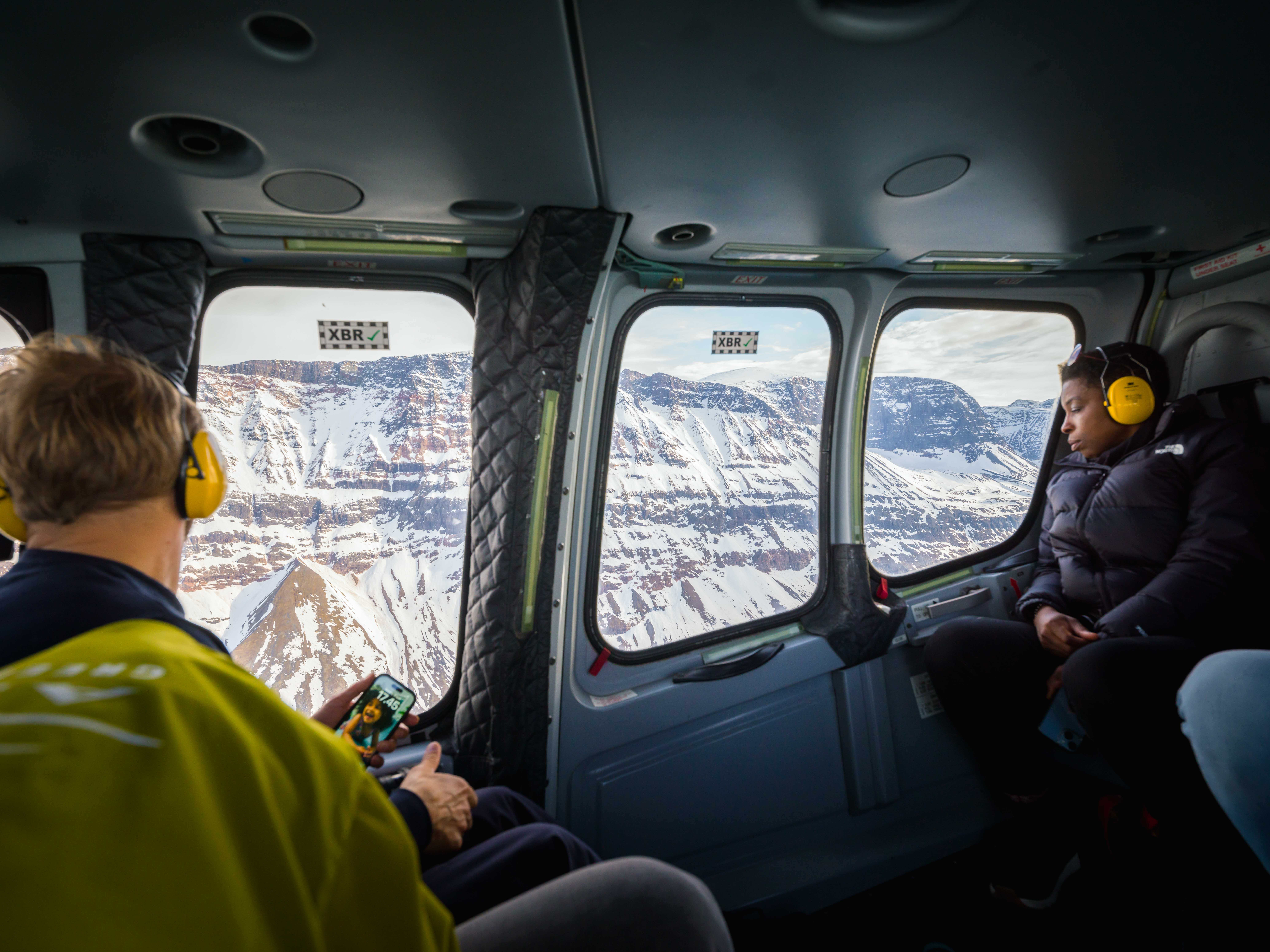 A woman wearing yellow ear protection and a black jacket looks out an aircraft window at a snowy and rugged landscape.