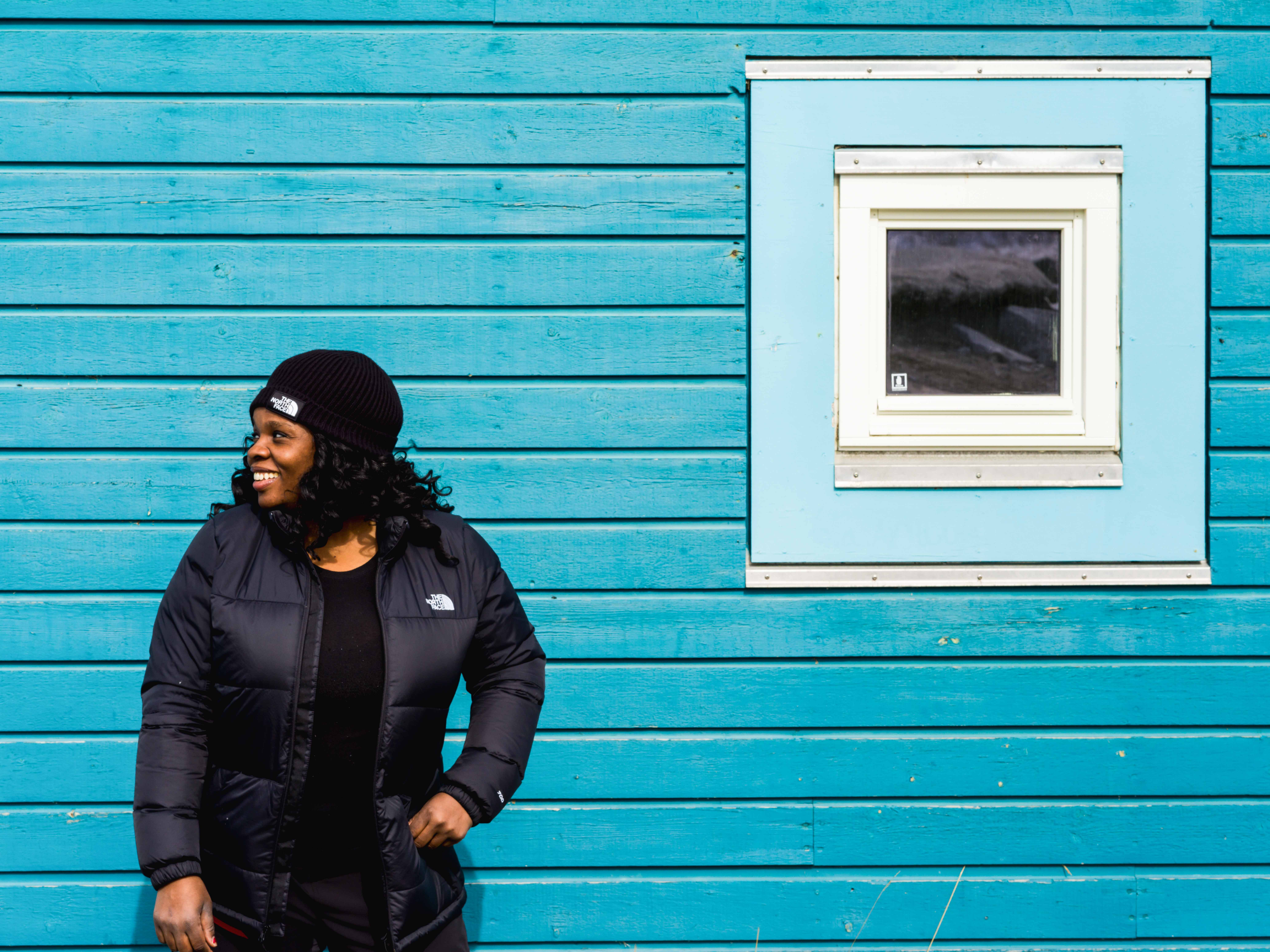 A Black woman in a black jacket and winter hat stands against the side of a building, which is painted in bright turquoise.