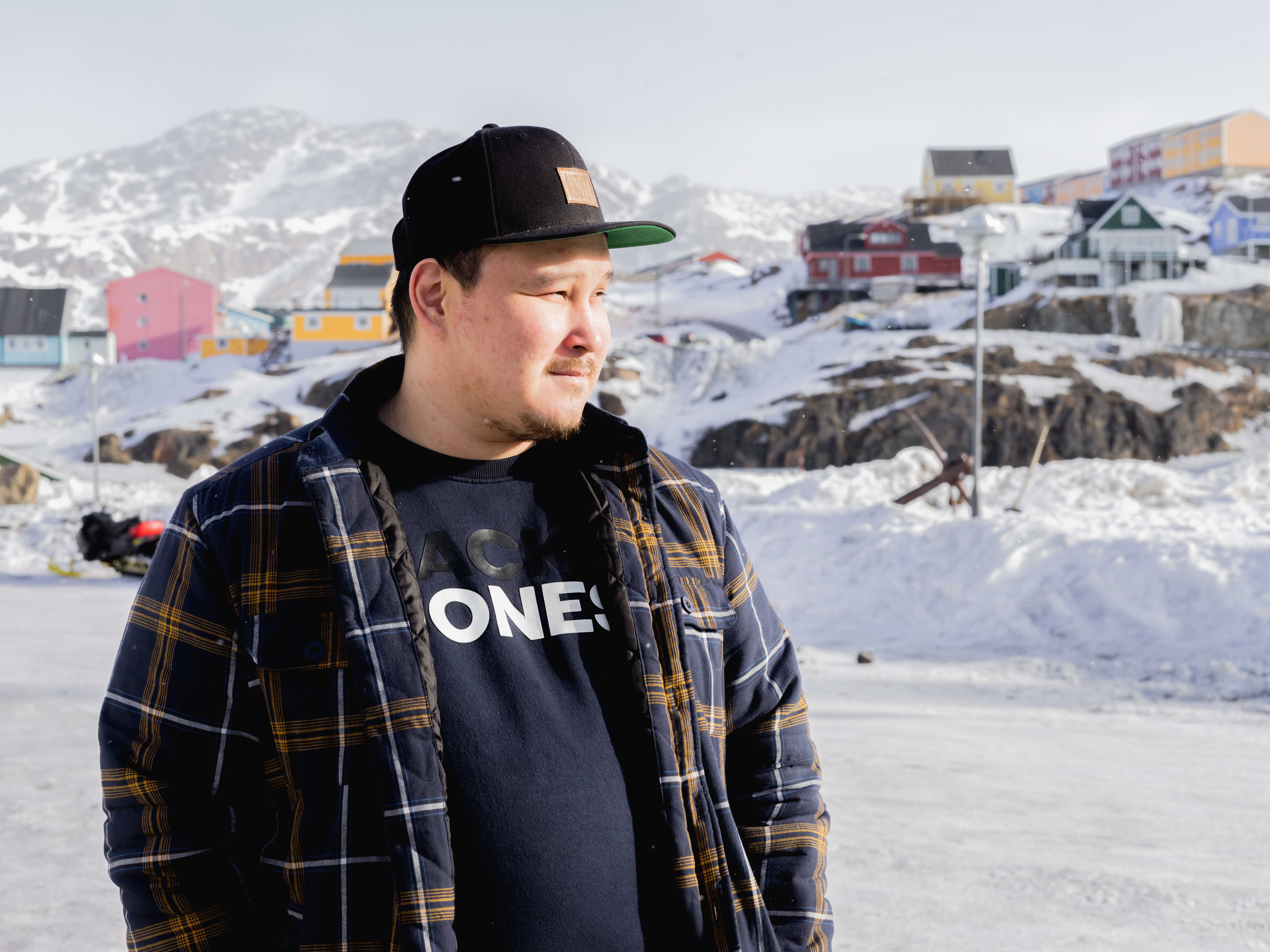A man in a black cap and blue and orange plaid jacket looks to the right of the frame. Behind him. a snow covered town of colorful buildings is visible.