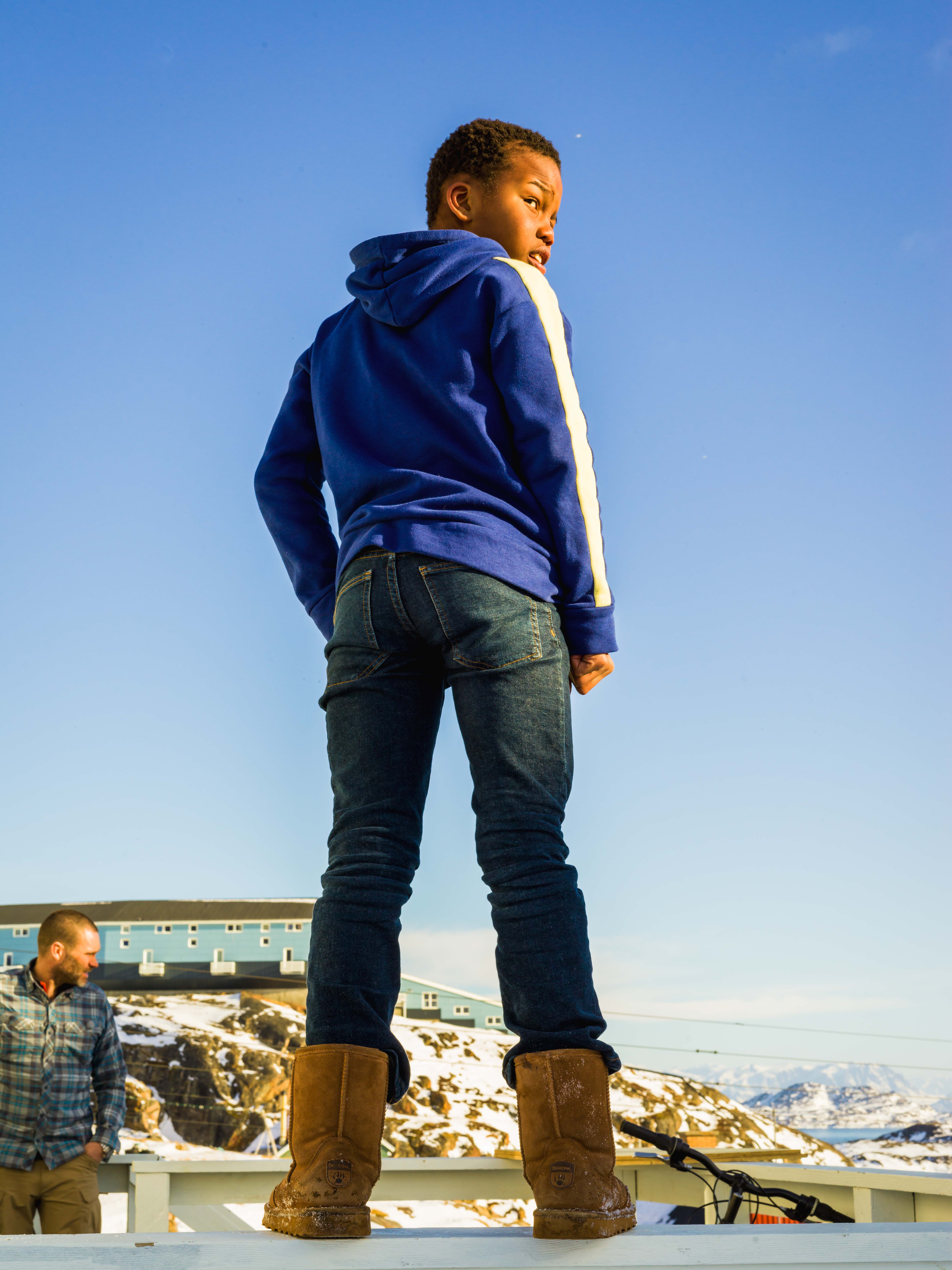 A young boy stands facing away from the camera and gazes over his shoulder. He has dark skin and wears a blue hoodie and jeans.