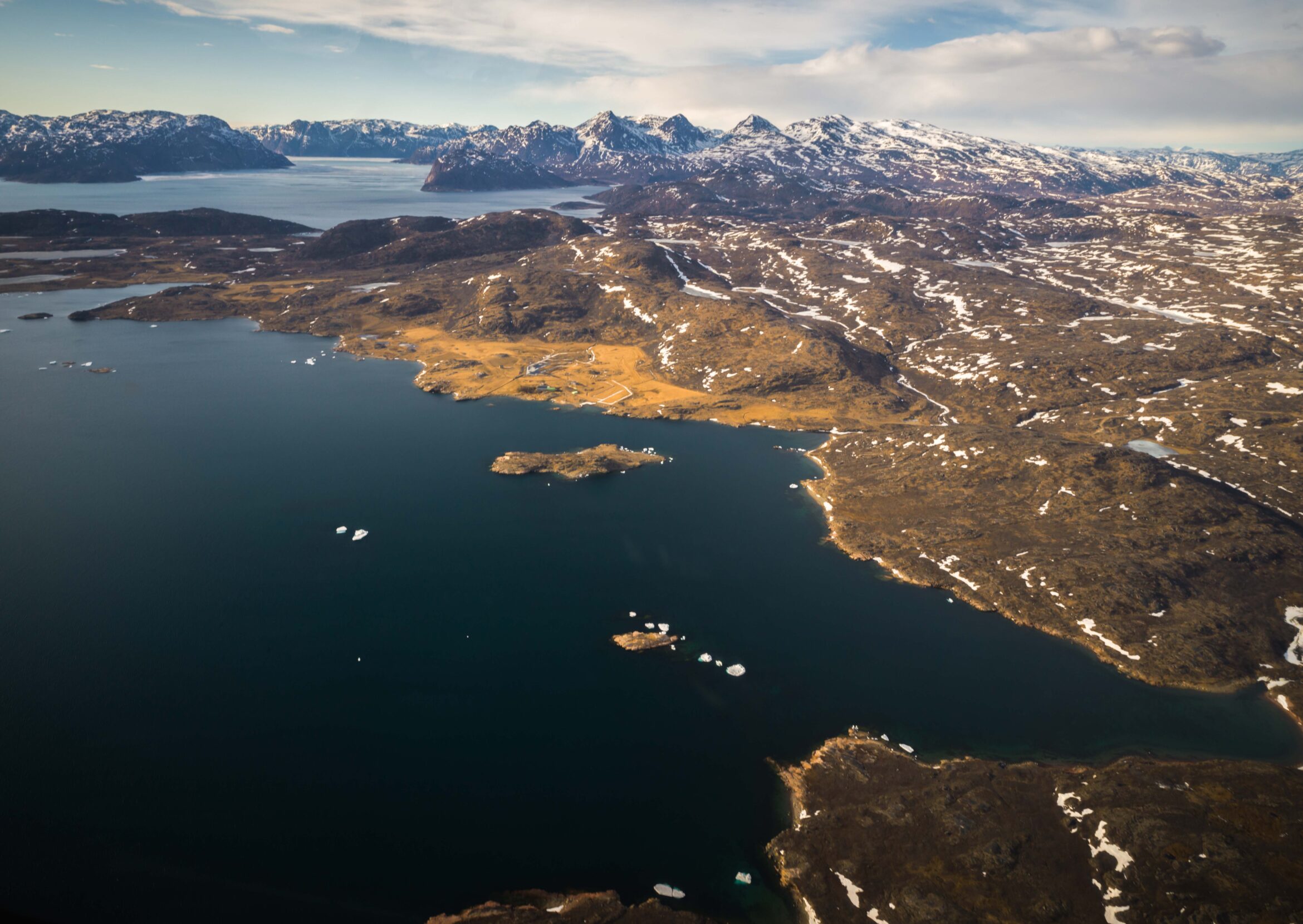 Amber mountains rise from the blue waters of a fjord. Patches of snow dot the land.