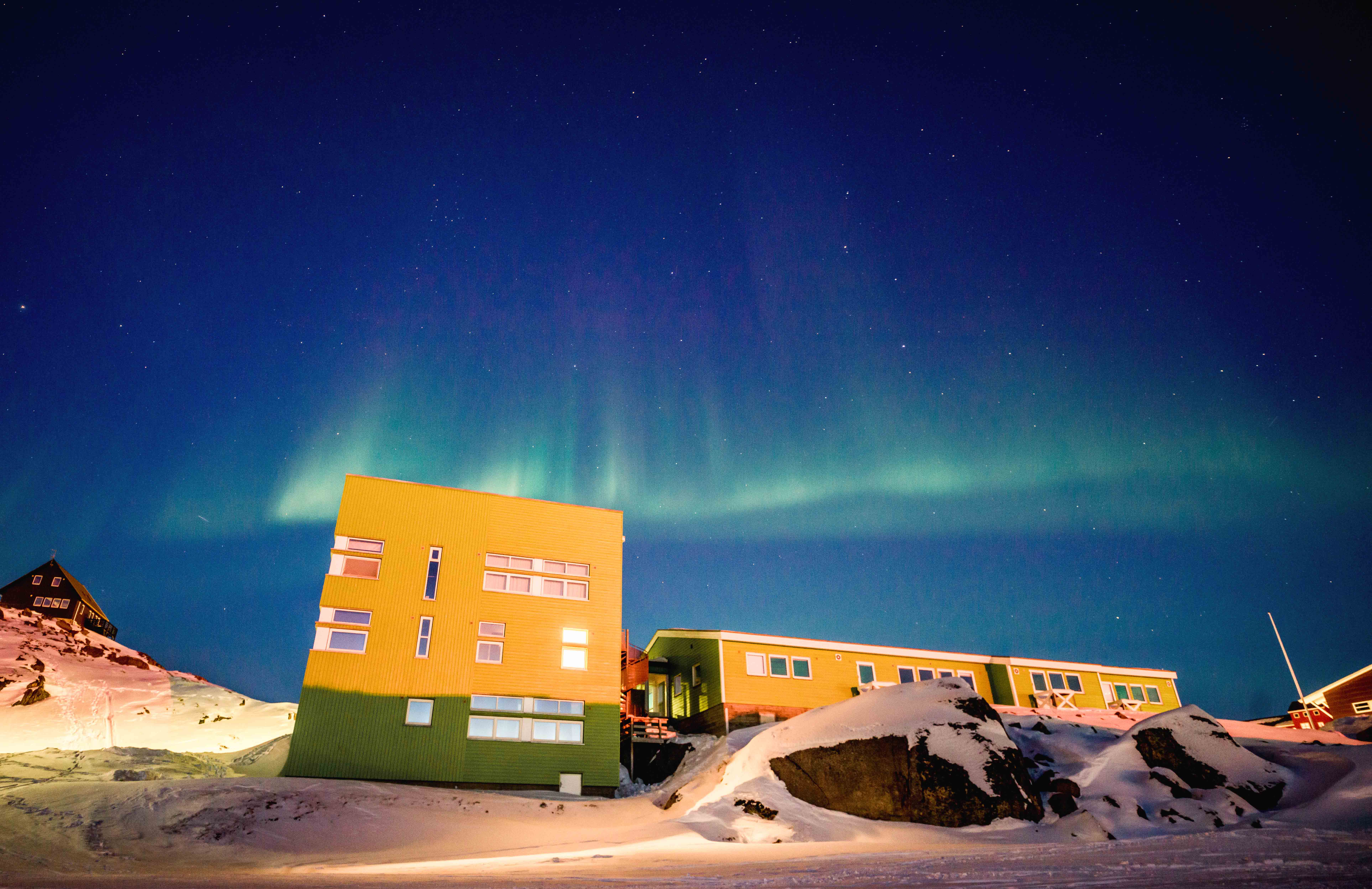 Above a green and yellow building and a snow covered street. A green band of aurora arcs across a blue night sky.