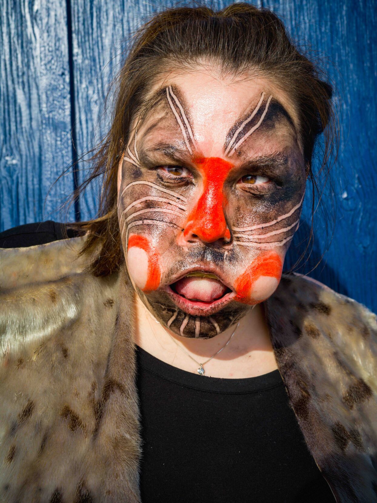 A woman stands against a blue-painted wooden backdrop. Her face is painted in black and red and her cheeks puff out.