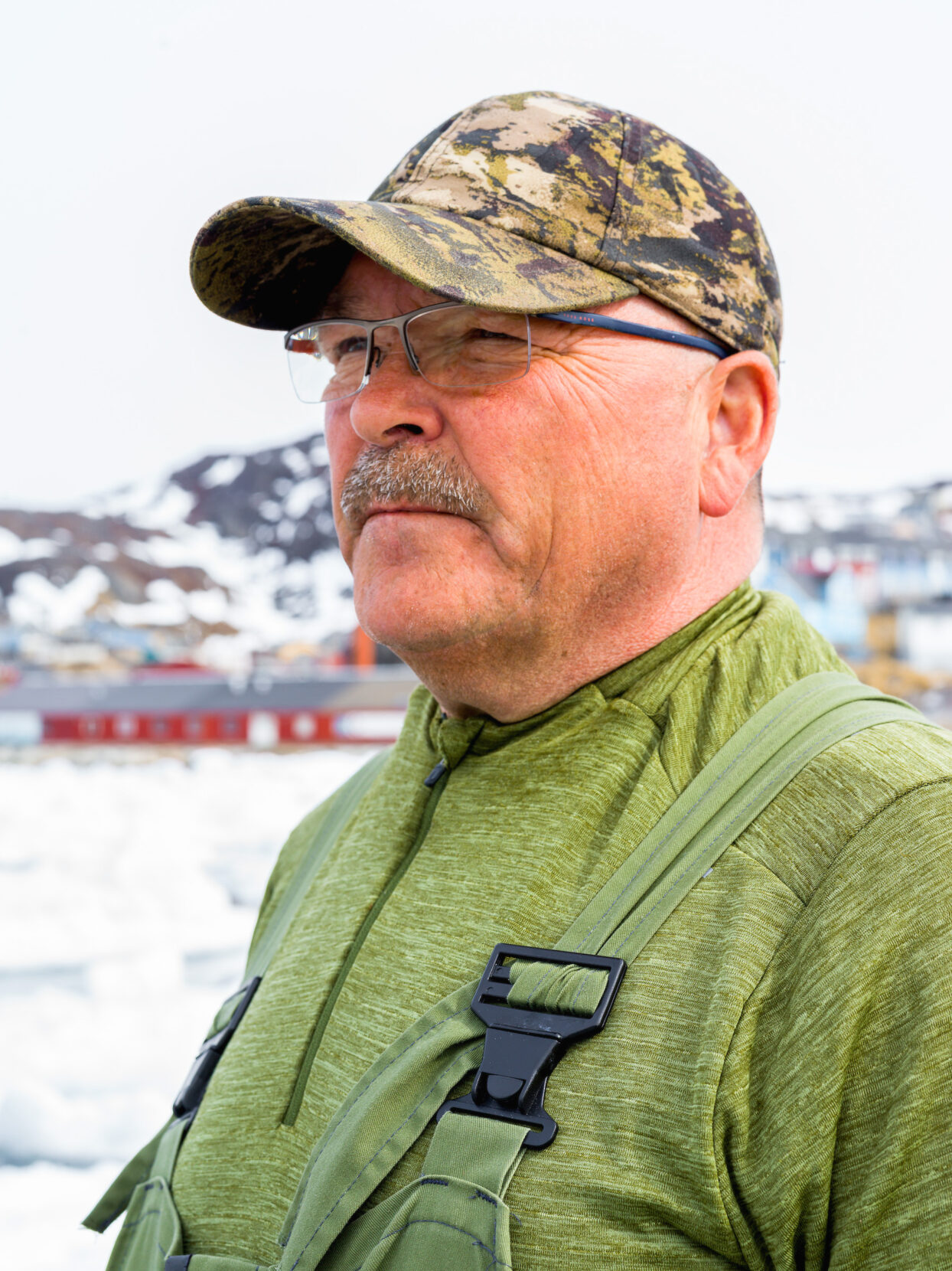 Portrait of a man with a mustache, glasses, and camouflage cap staring into the distance, with a snowy landscape and icy waters behind him.