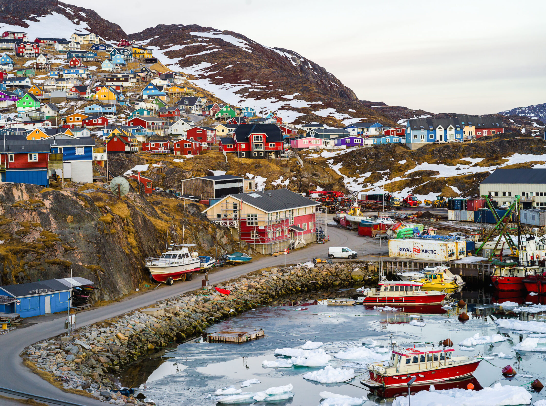 A drone shot of a rocky, coastal town of brightly colored buildings and boats with snow and ice.