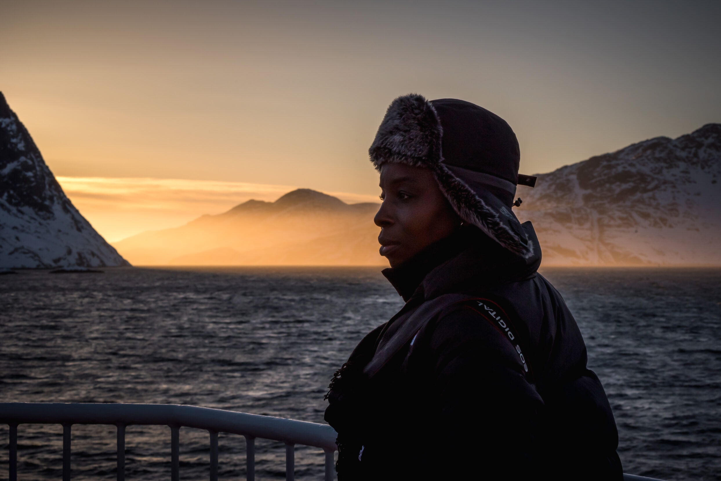 A black woman in a fuzzy hat stands at a boat railing in front of a glowing horizon of snowy mountains and silvery waters.