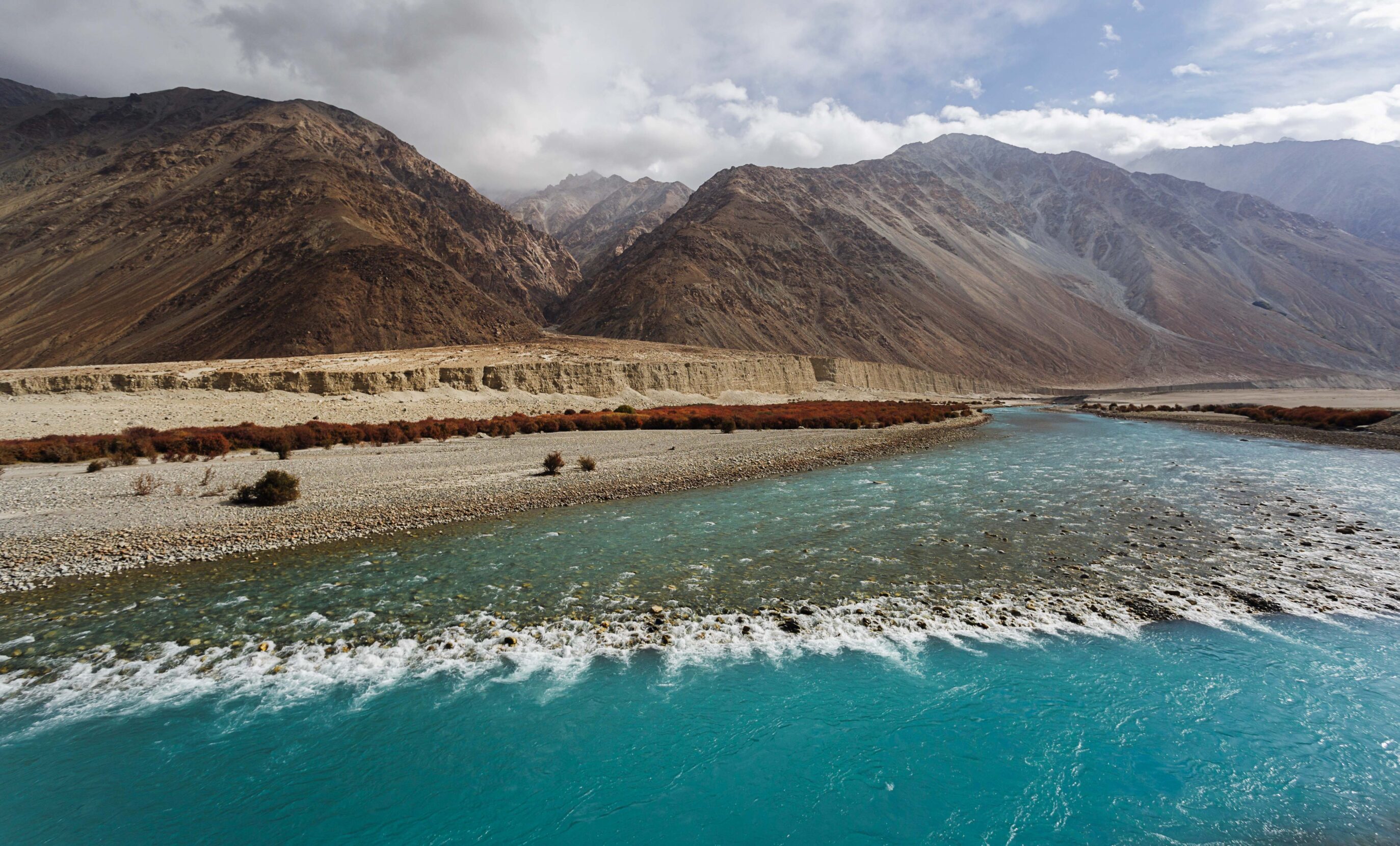 A stark, treeless mountain range stands behind the wide turquoise waters of the Indus river.