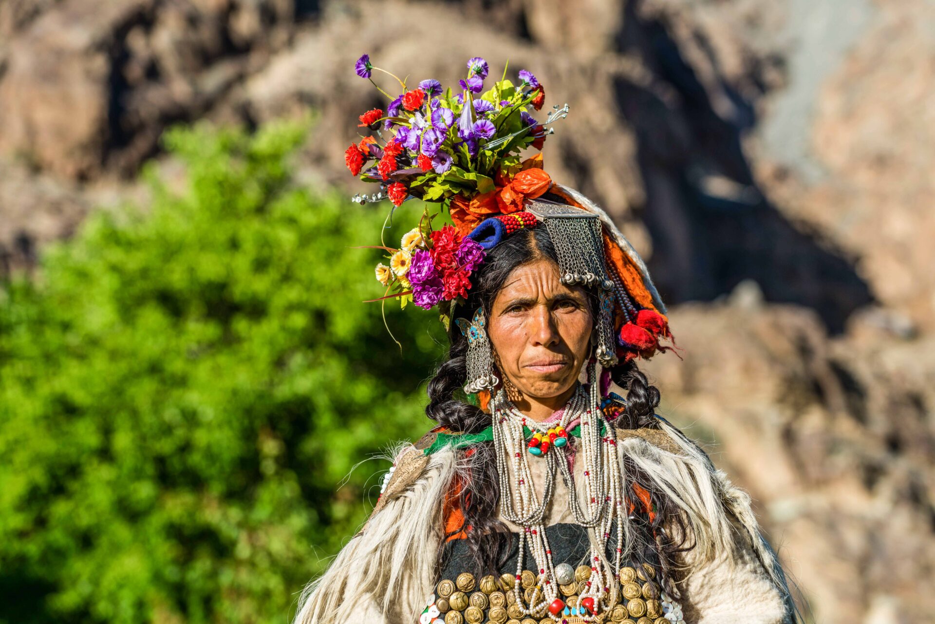 Portrait of a woman in a colorful Brokpa headdress topped with purple and red flowers.