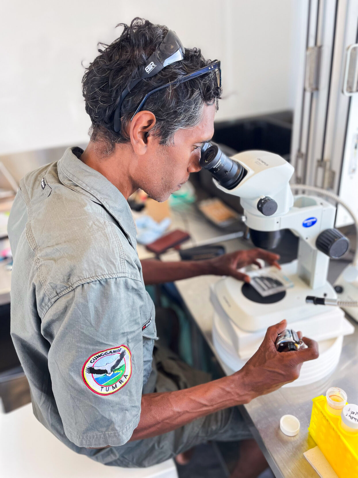 An Indigenous Australian man peers through a microscope. His jacket bears a patch that reads 