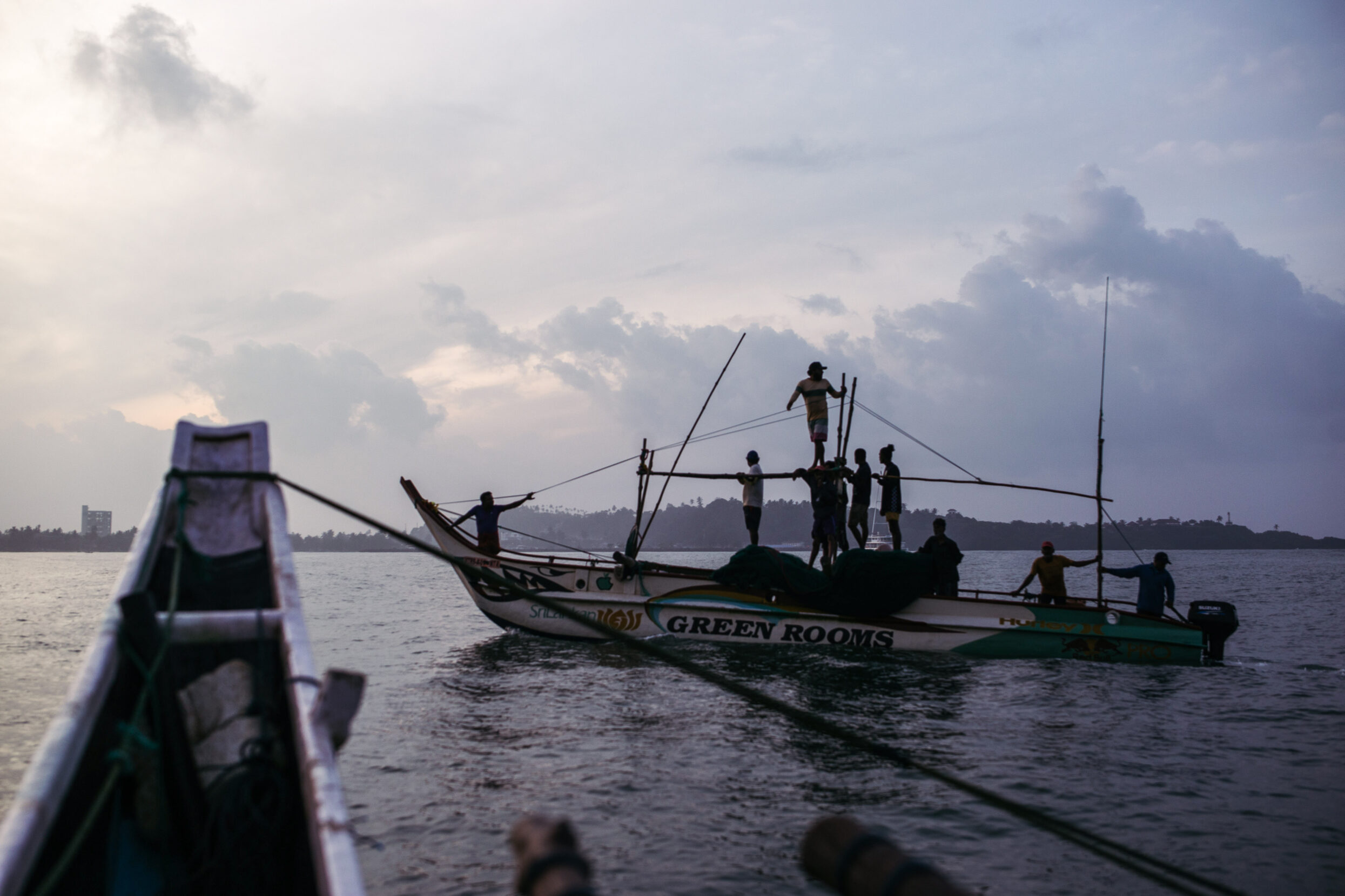 A boat with silhouetted fishermen balancing on poles appears before the bow of another boat.