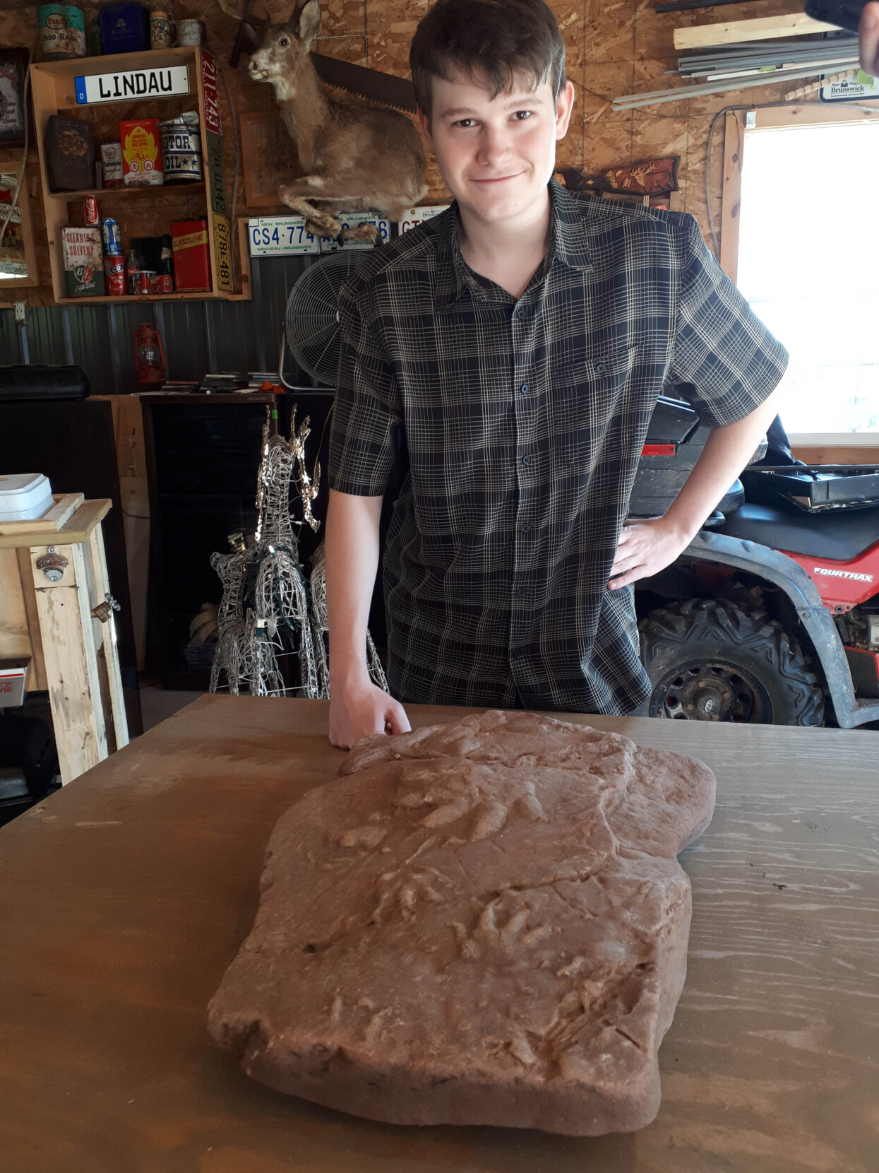 A teenage boy in a checkered shirt smiles with his hand on his hip. In front of him sits a two-foot slab of red rock with fossilized footprints on it.