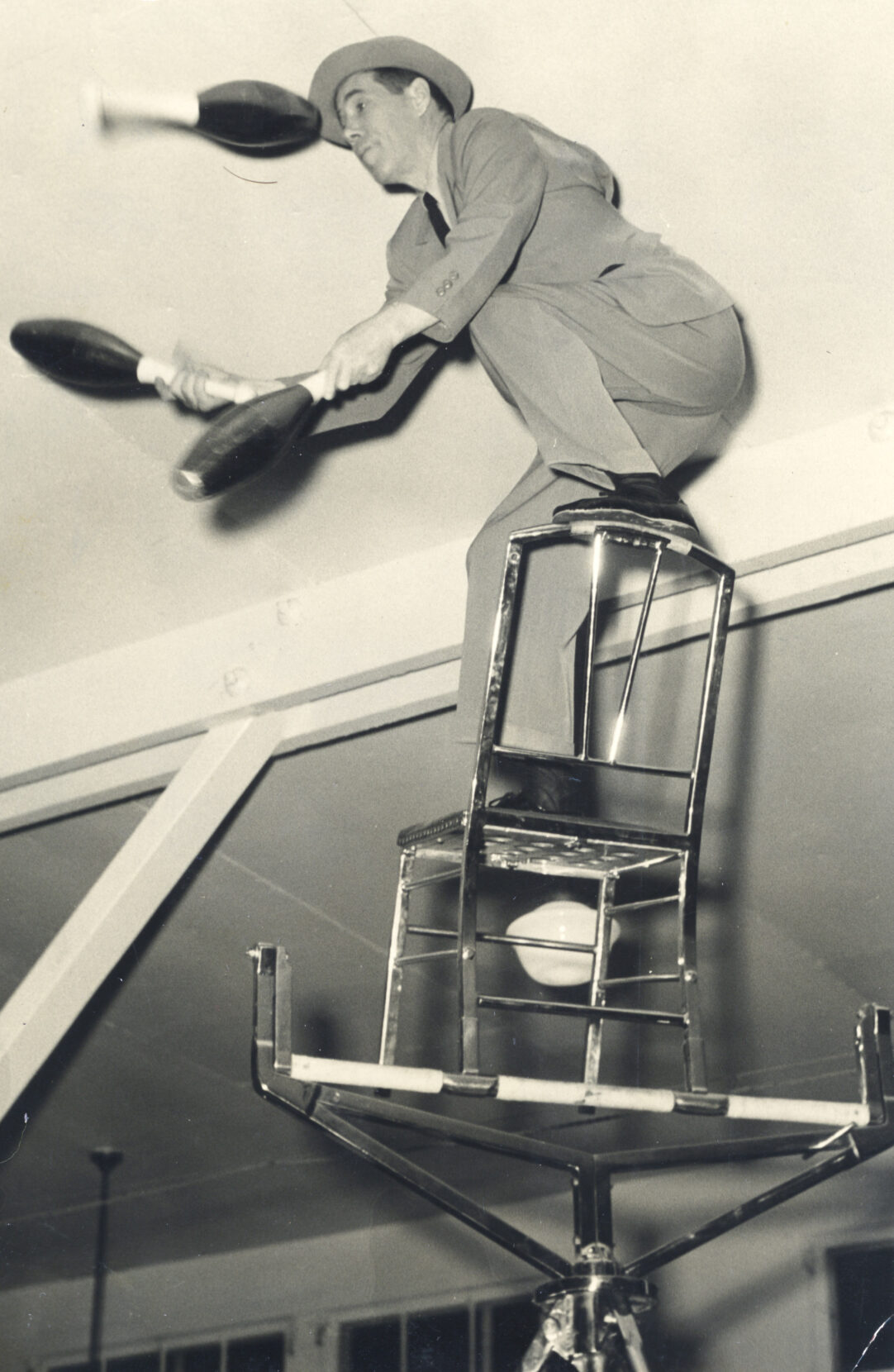 Black and white photo of a man in a suit, balancing on a chair while juggling.