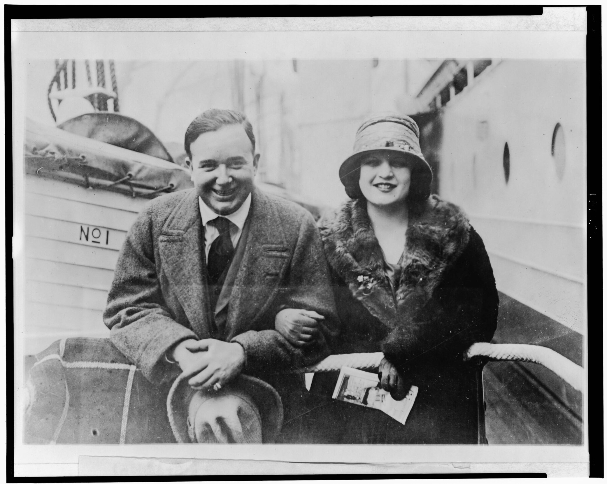 Black and white portrait of a man and woman, arms linked, before a ship at dock.