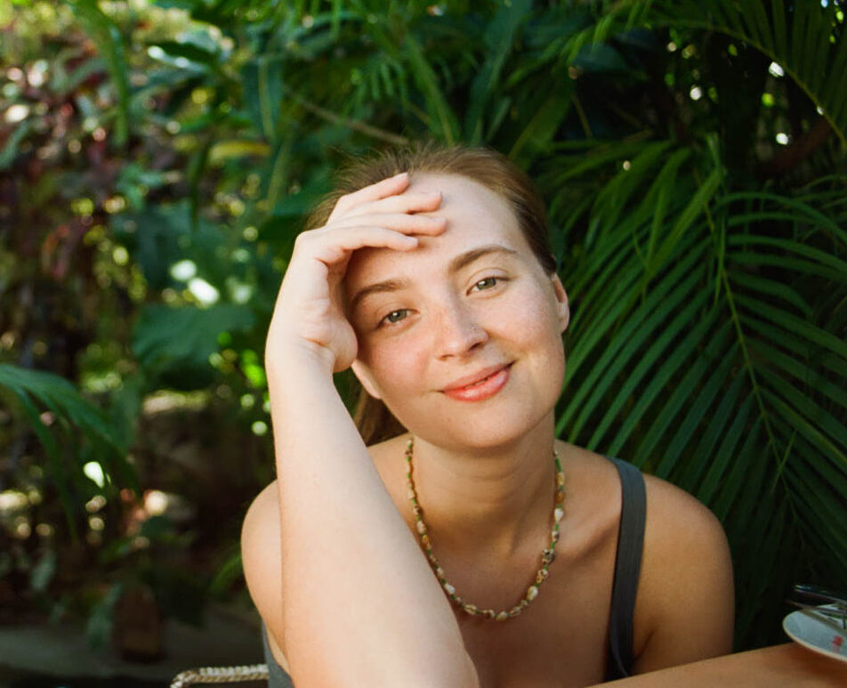 Portrait of a smiling young woman, seated and surrounded by tropical foliage.