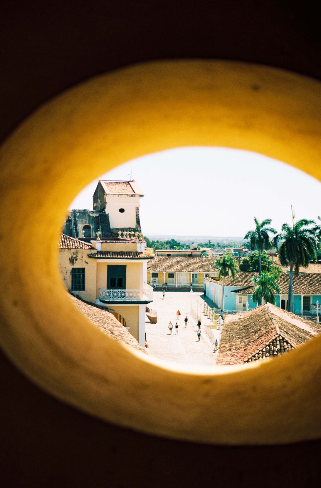 A peek through an oval wall opening of pedestrians walking amongst Spanish-tiled buildings and palm trees below.