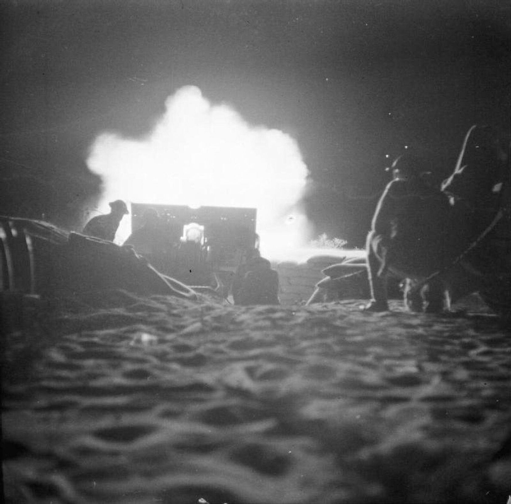 Black and white photo of silhouetted soldiers on the sand at night, with the plumes of an explosion on the horizon.