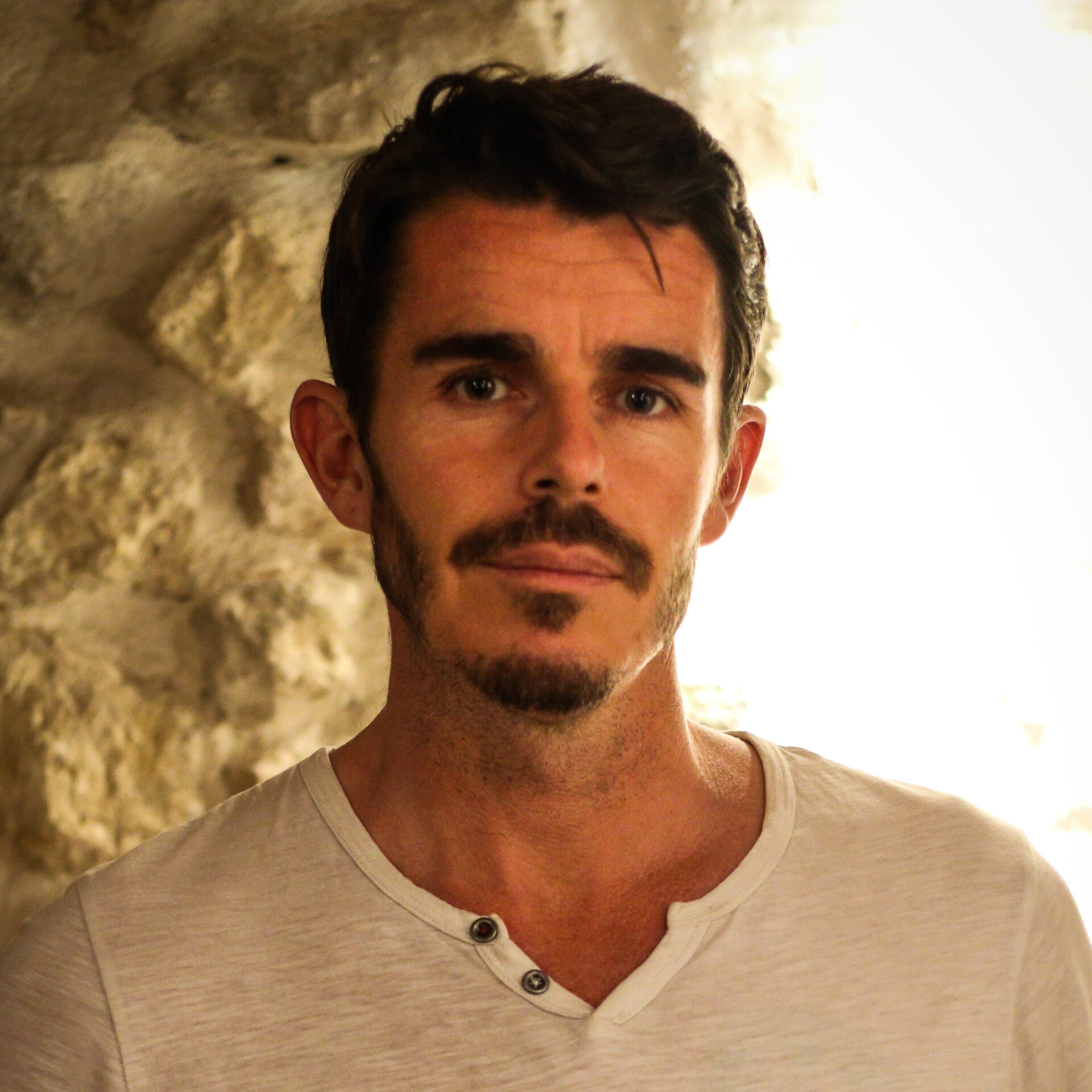 Portrait of a man in a white shirt with short brown hair, mustache and beard, standing before a rock wall.