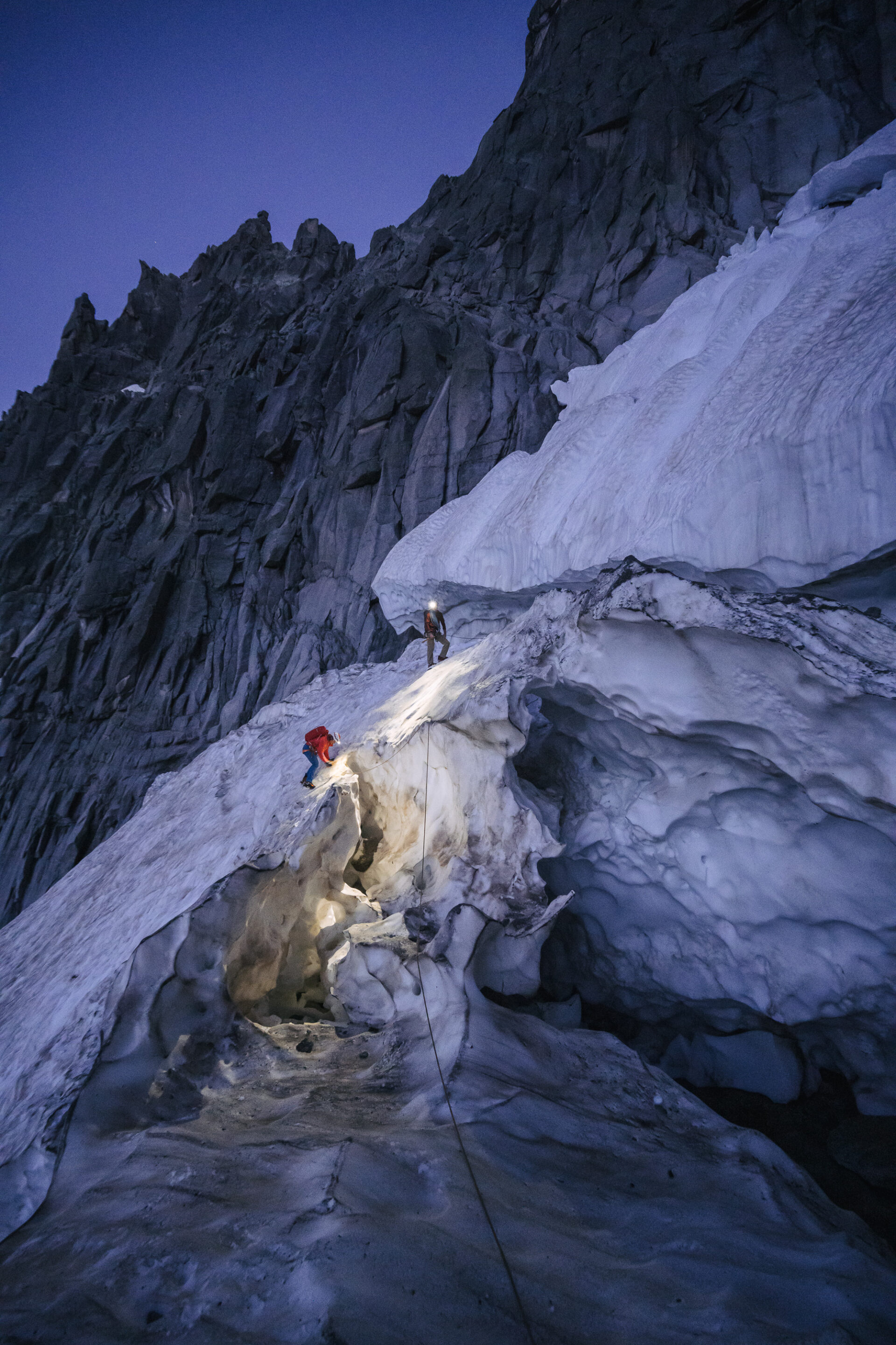 Two climbers on a snowy mountain side in the dark. The scene is illuminated by moonlight and their headlamps.