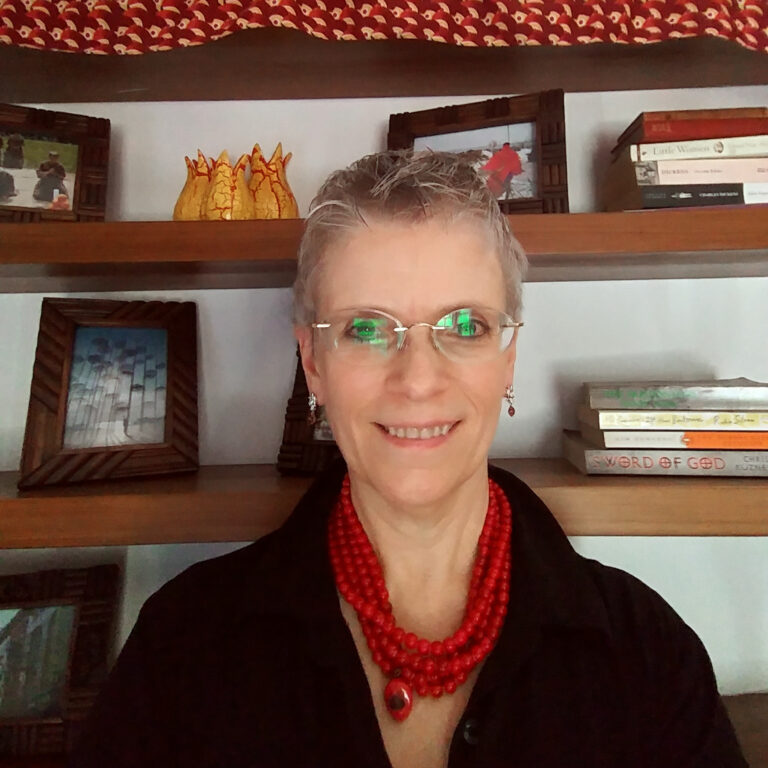 Headshot of a woman with short gray hair and glasses, wearing a red necklace and sitting in front of a bookshelf.