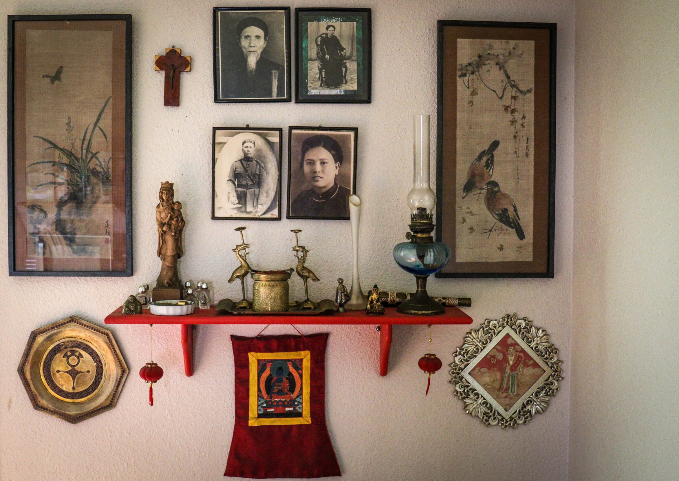 Four framed photos on a wall, with two pieces of art. There is a red shelf on the wall with statues of religious ssymbols.