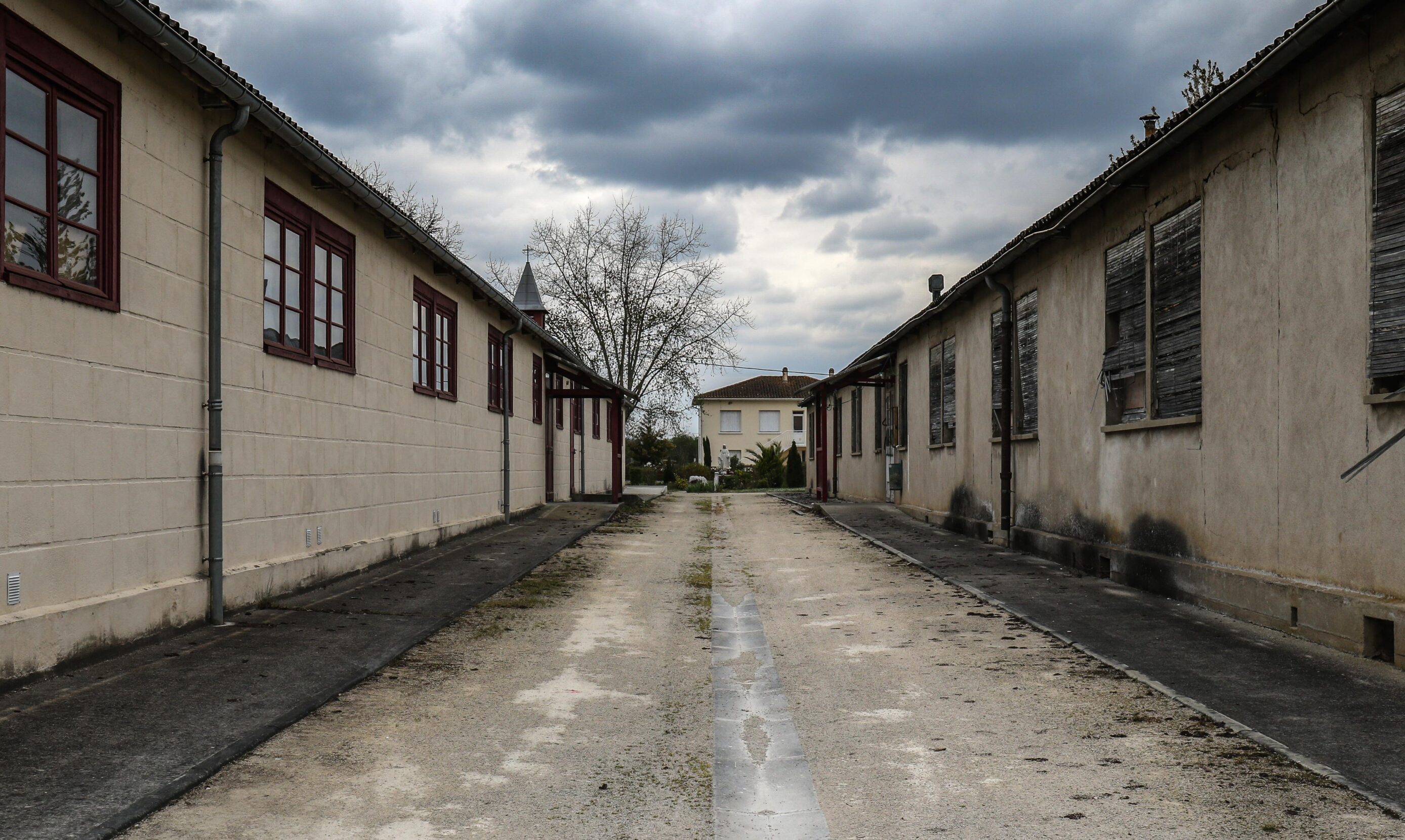 An empty alley between two buildings, one with windows painted with red trim, the other with windows covered by disintegrating blinds.