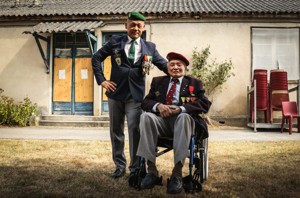 Two older men with military medals. One stands in a green beret and matching tie with a chest full of medals. The other sits in a wheel chair, with a red beret and fewer medals.