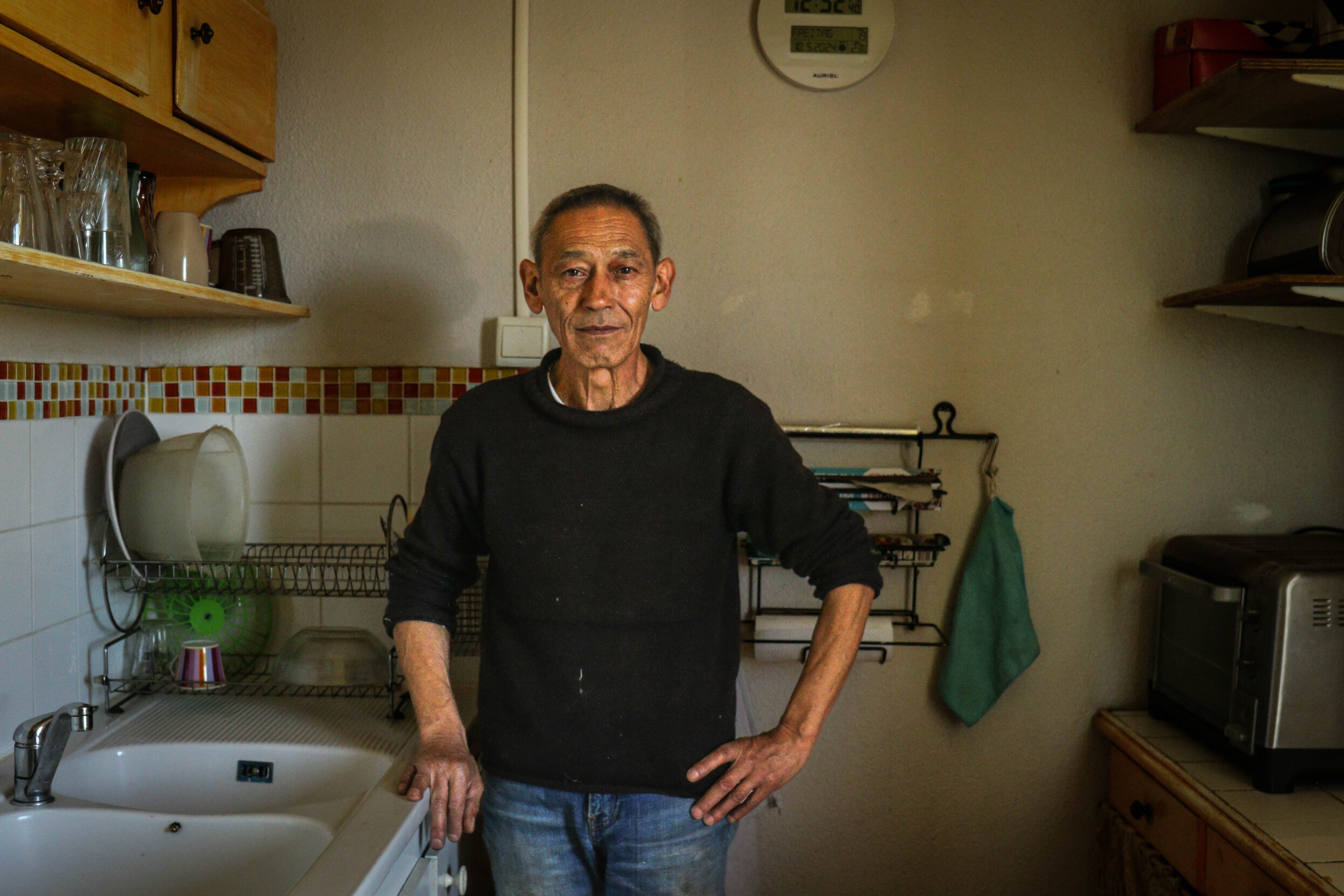 An older man with dark gray hair and a black sweater stands in a kitchen.