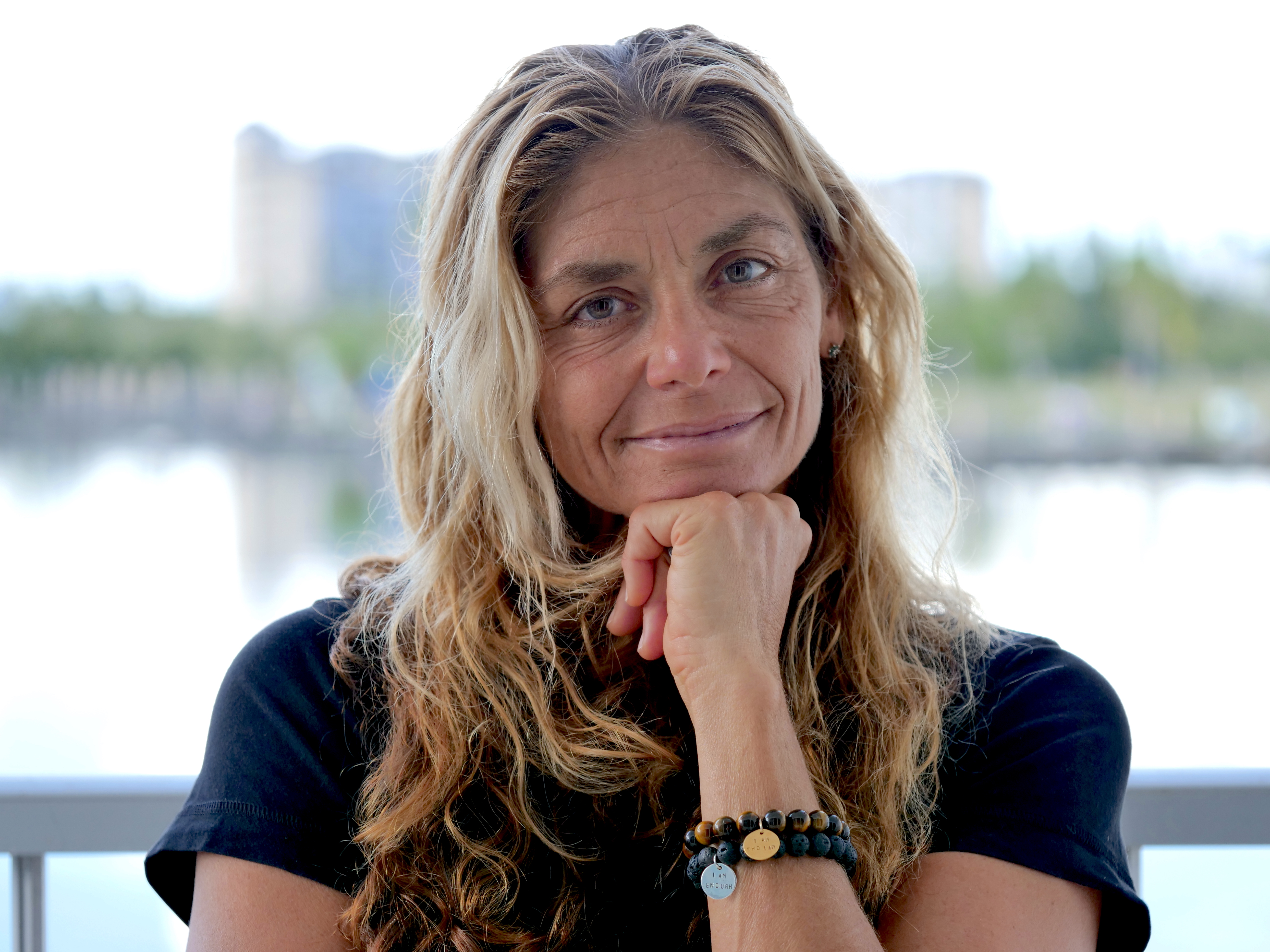 Portrait of a woman with long, curly, blonde hair, in a navy shirt, with a river and urban landscape in the background.