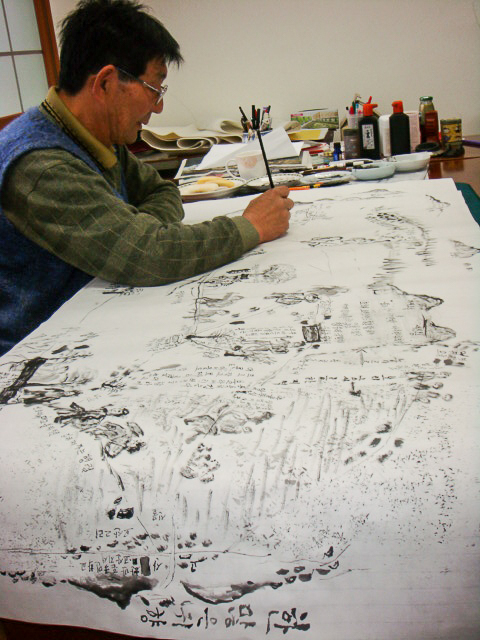 An older Asian man wearing glasses uses a long pen to draw on a large sheet of paper.