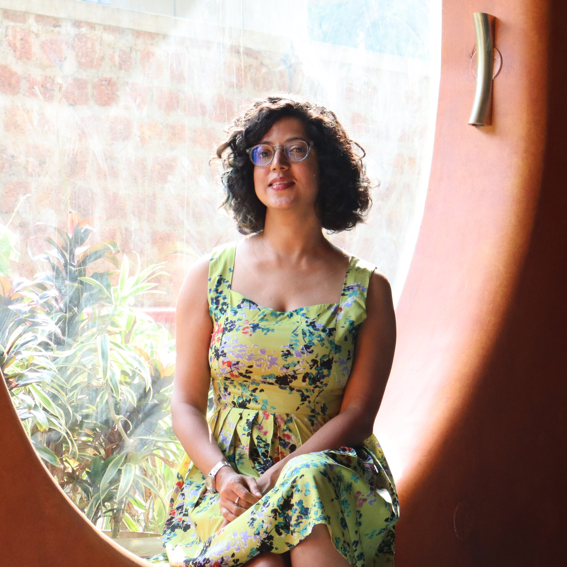 A woman in a green dress and glasses sits by an oval window.