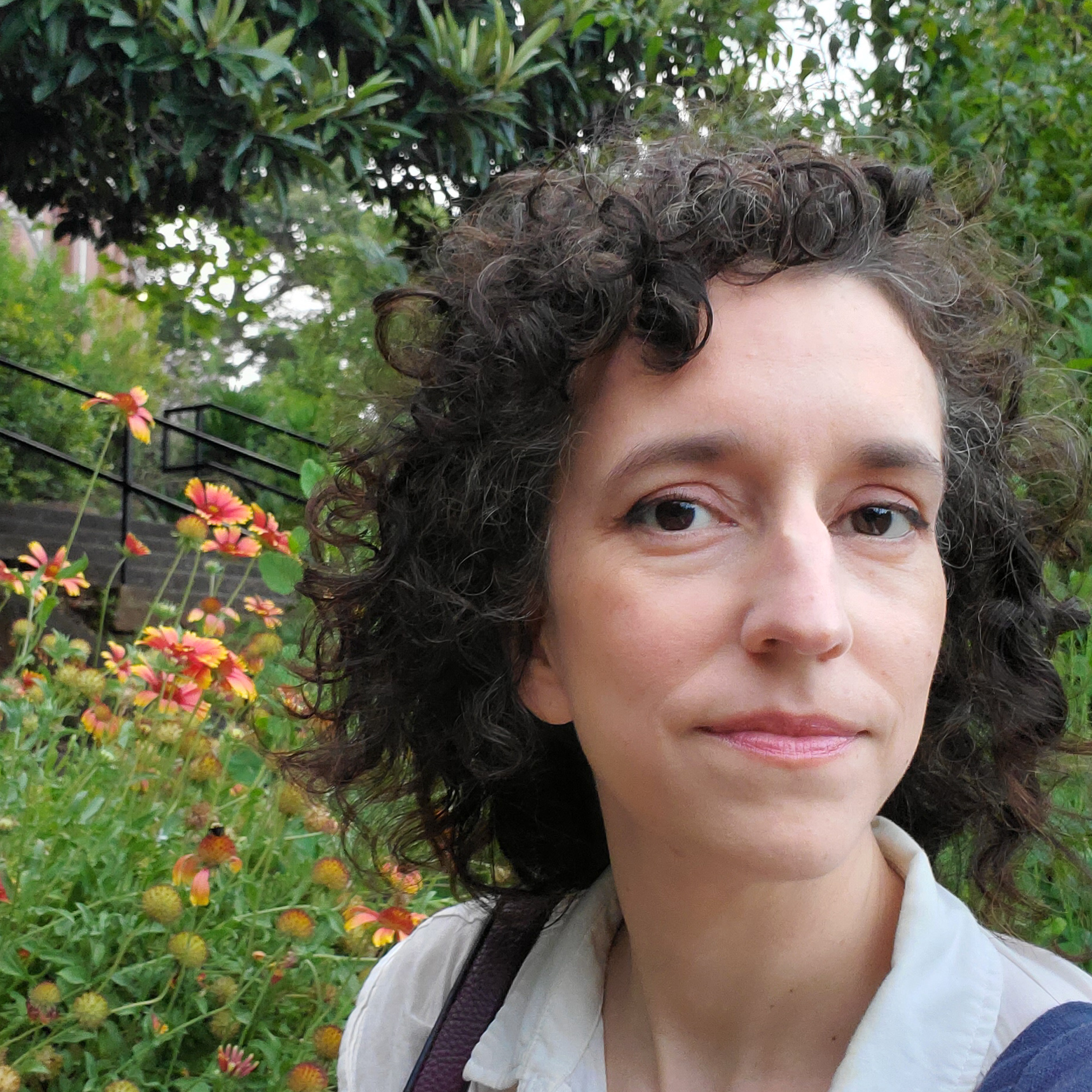 Portrait of a woman with dark curly hair standing alongside orange flowers.