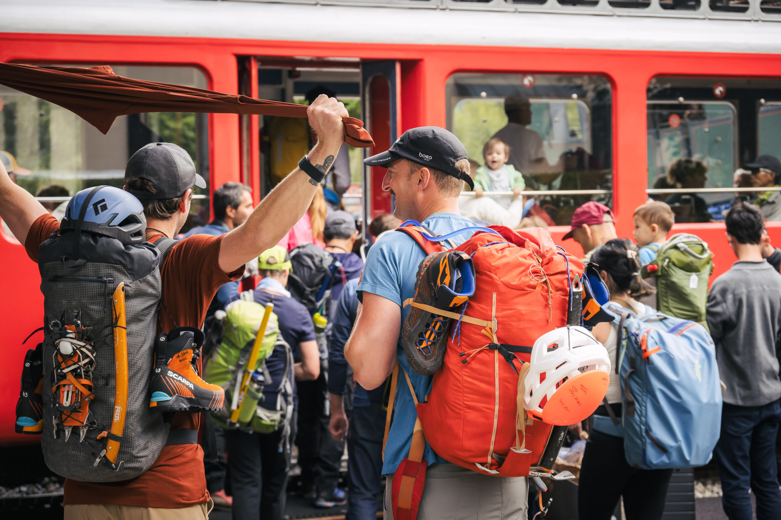 A large group of people waits to board a red and white train.