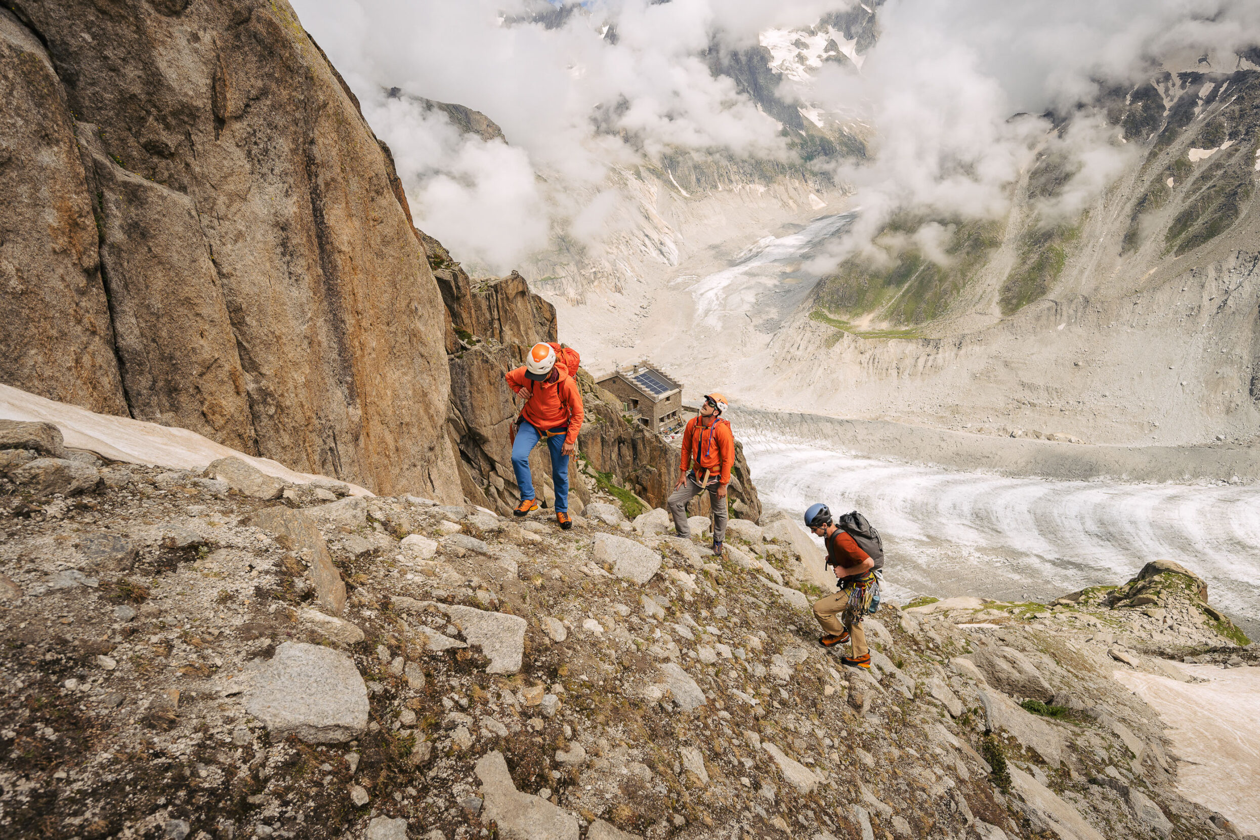 Three people on the side of a mountain with a small building in the distance perched on a cliff.