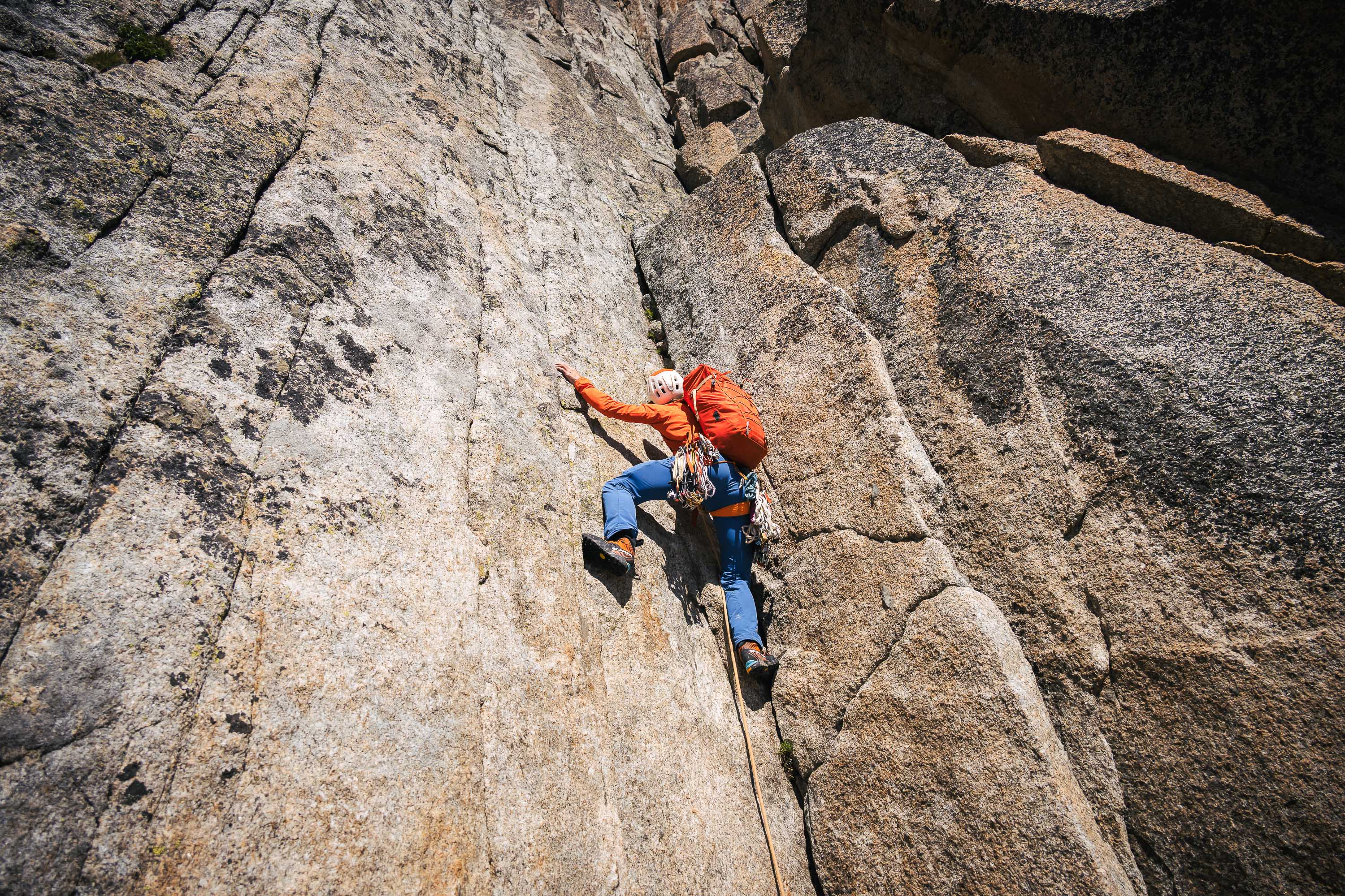 A climber in blue pants and an orange jacket scales a rock face.