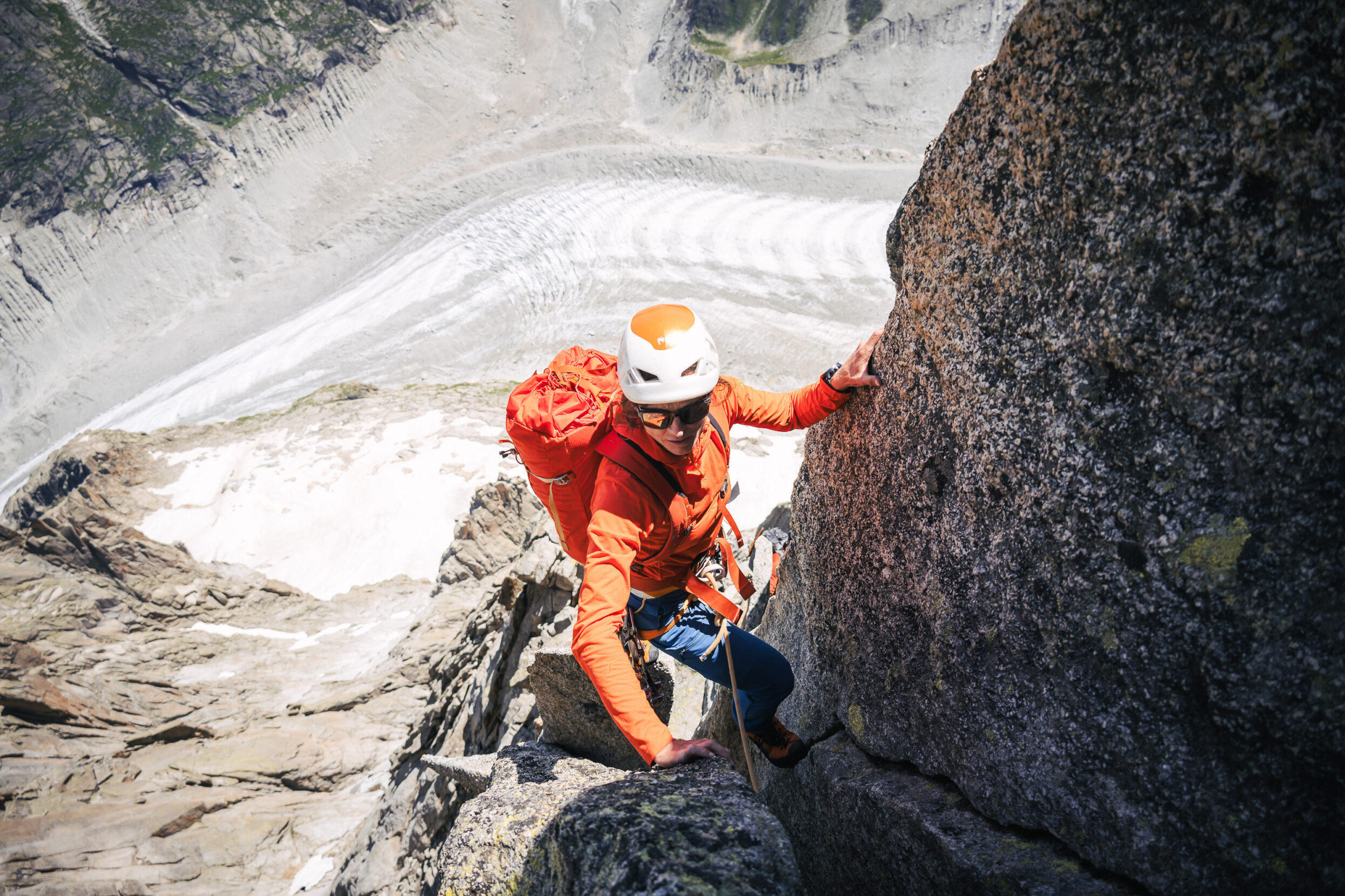 A female climber in an orange jacket perched high above a glacier.