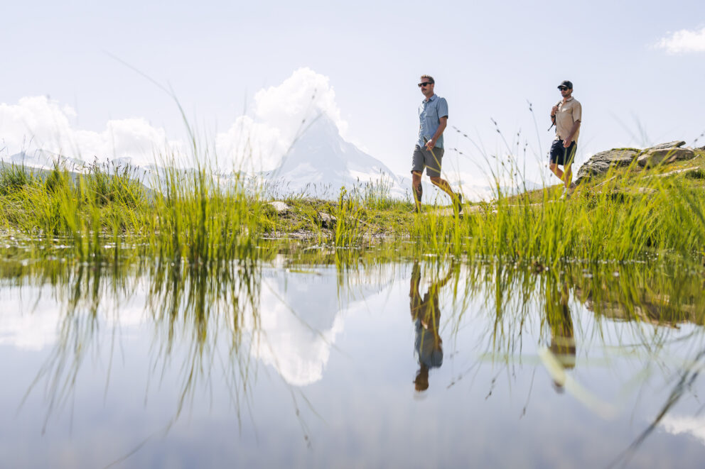 two men walk reflected in a lake along with a mountain.